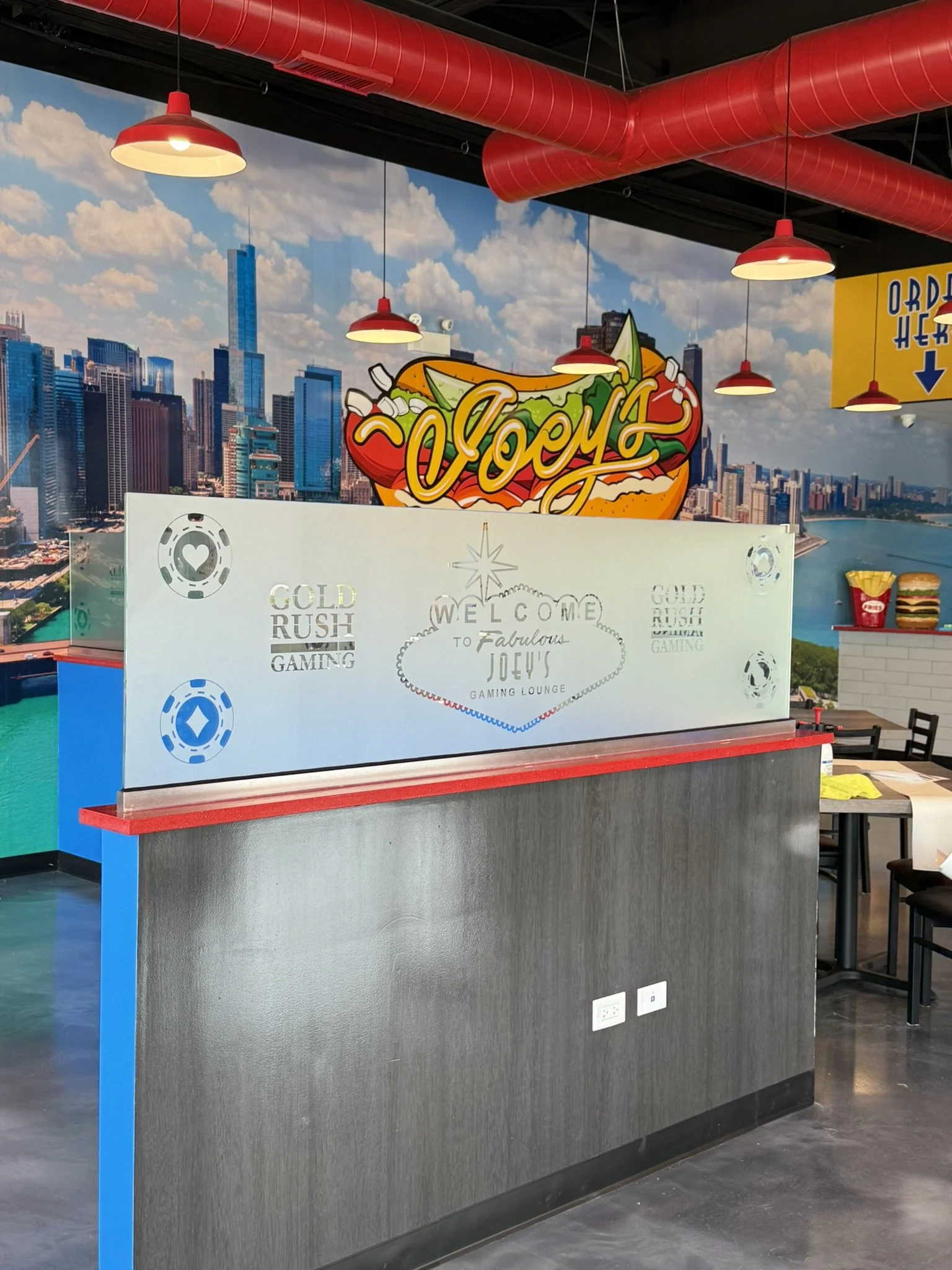 Indoor space with a mural of skyscrapers, red pipes and hanging lights, decorated with a hot dog graphic that says 'Enjoy'. There is a partial white divider with gaming-related graphics and a welcome message.