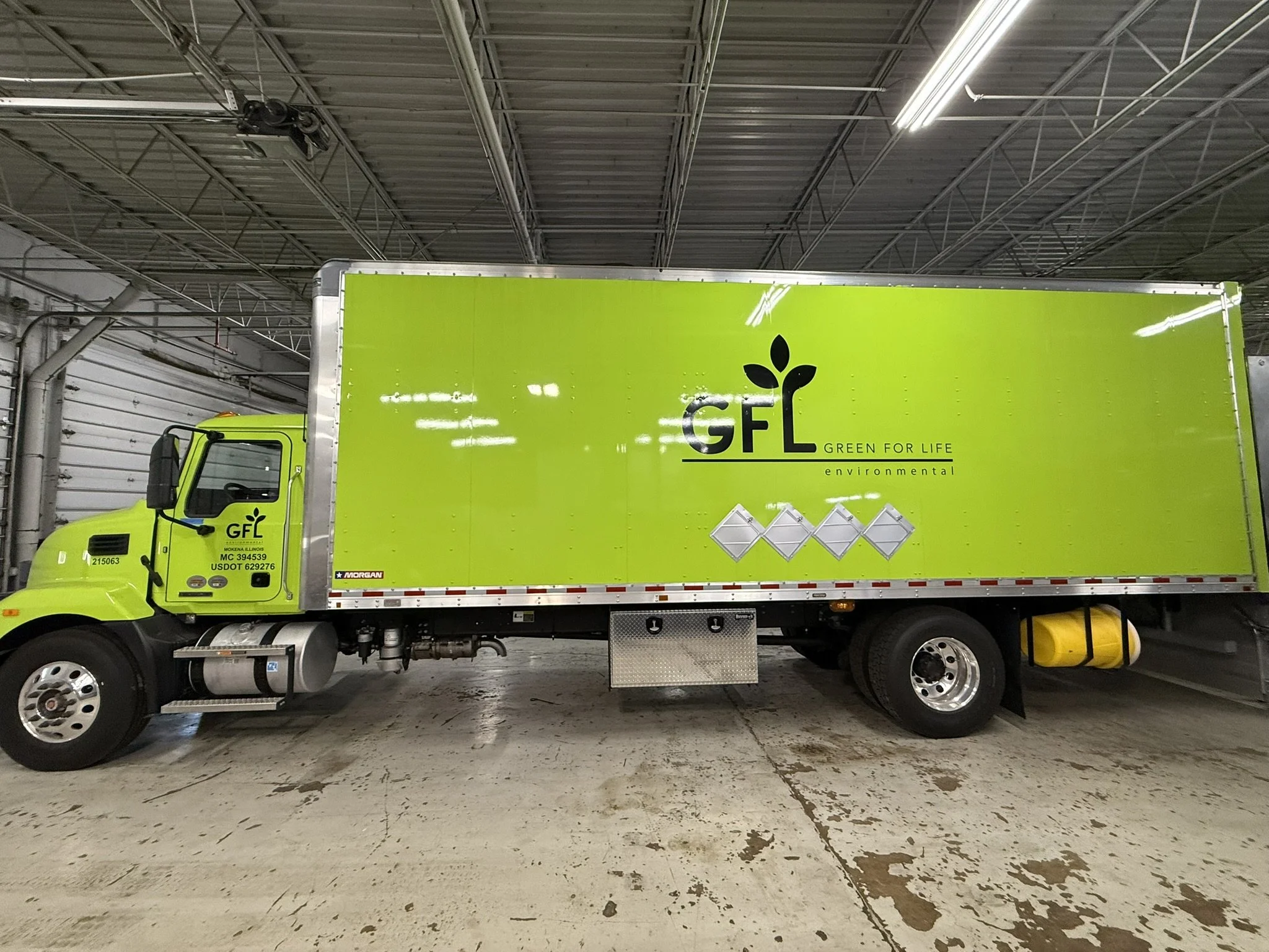 Bright green delivery truck with 'GFL Green for Life' logo parked inside an industrial warehouse with a metal roof and concrete floor.