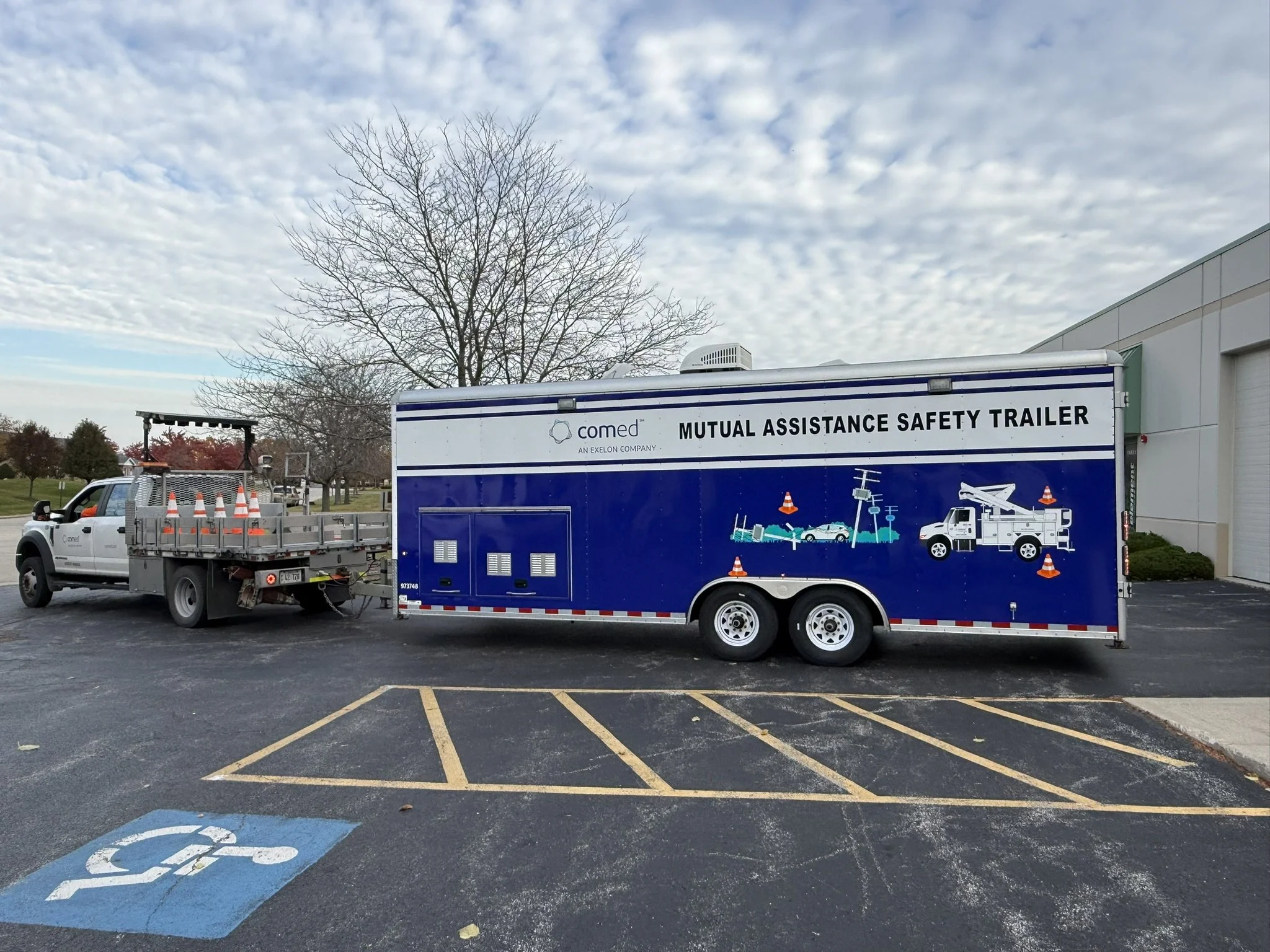 A parking lot with a blue trailer labeled 'Mutual Assistance Safety Trailer' parked next to a white utility truck. The lot has a blue handicap parking space and yellow striped parking spots. There is a leafless tree and a building in the background u