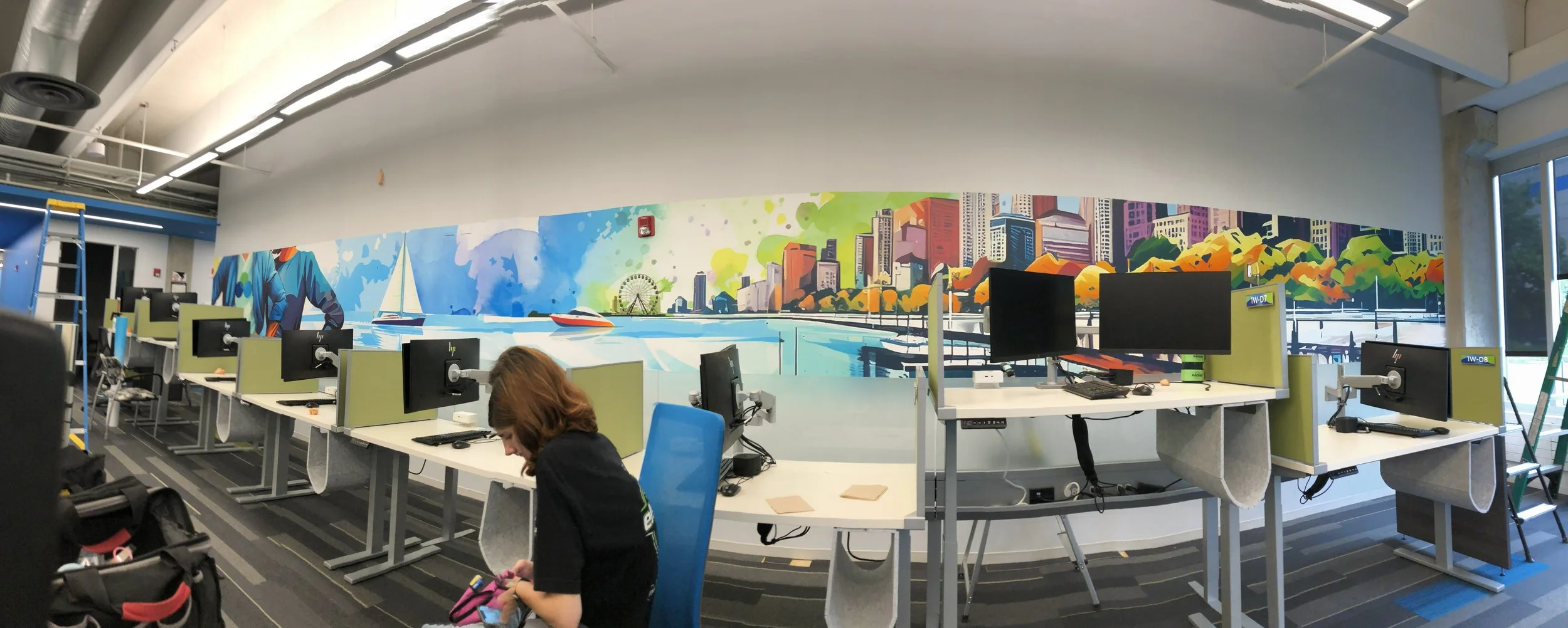 Office space with multiple desks, monitors, and dividers. A colorful cityscape mural on the wall features a skyline, sailboat, water, and a Ferris wheel. An woman with brown hair is seated in the foreground, looking at her phone. There is a ladder an