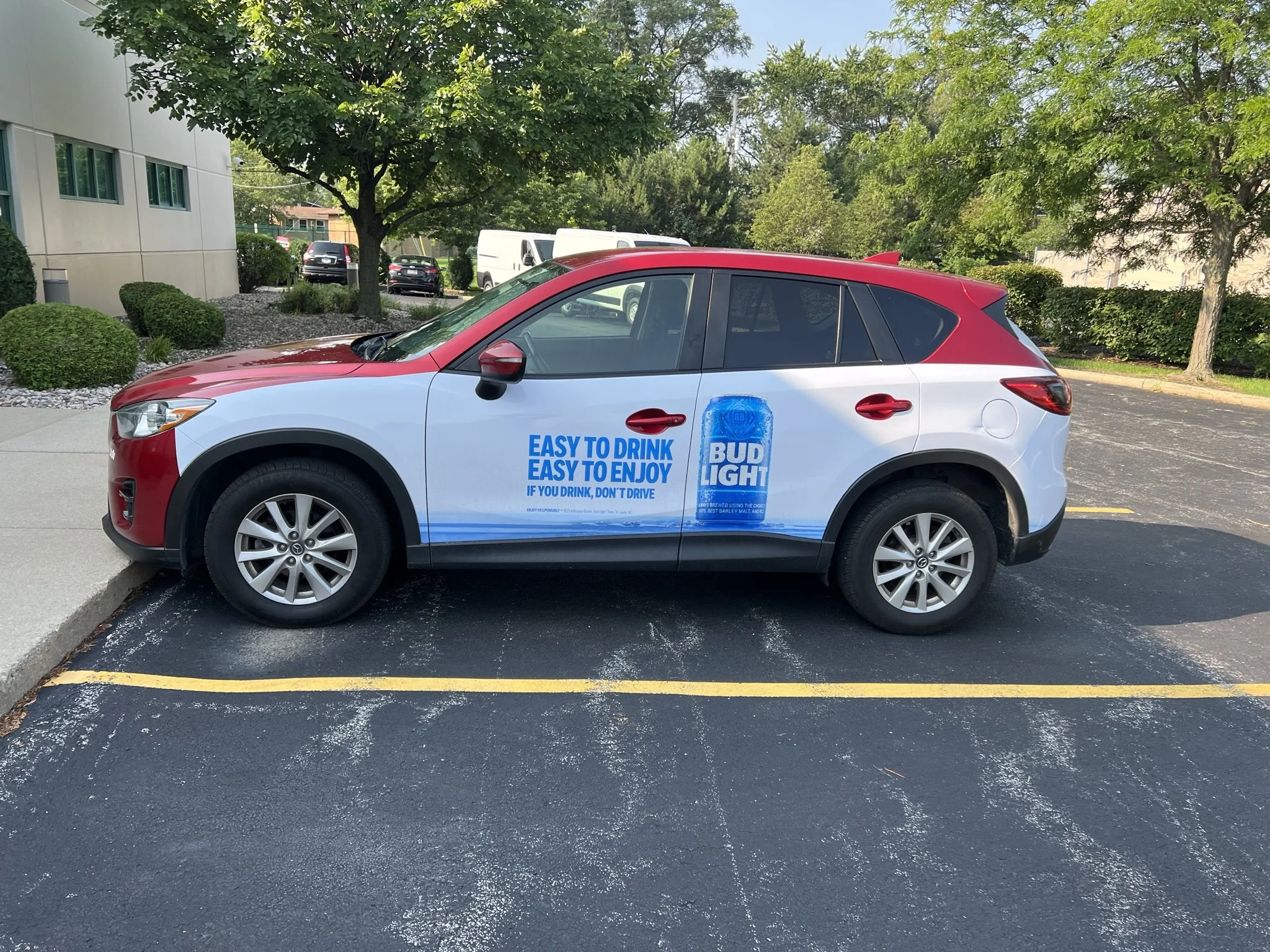 A white and red SUV parked in a parking lot next to a sidewalk, with trees and bushes in the background and a building on the left side of the image. The vehicle has an advertisement on its side promoting Bud Light beer.