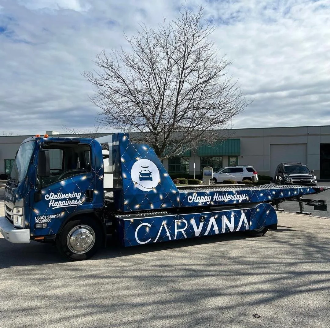 A blue car carrier truck with the Carvana logo and slogan, 'Delivering Happiness,' parked on a street in front of a parking lot and warehouse buildings, with a leafless tree and cloudy sky in the background.