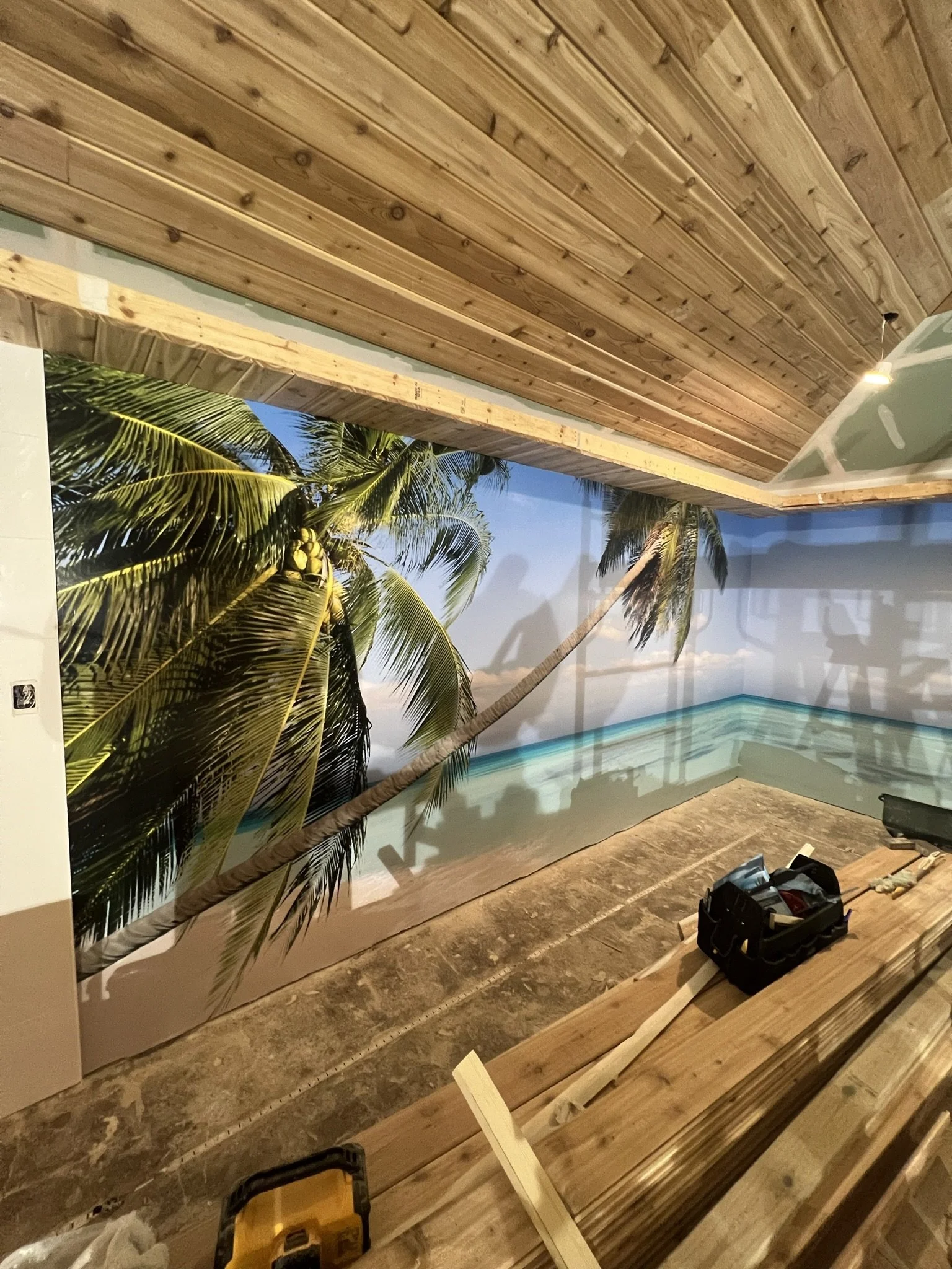Interior space under construction with a wall mural of palm trees, a swimming pool, and a bright blue sky. The ceiling has wood paneling, and there are construction tools and materials on the floor.