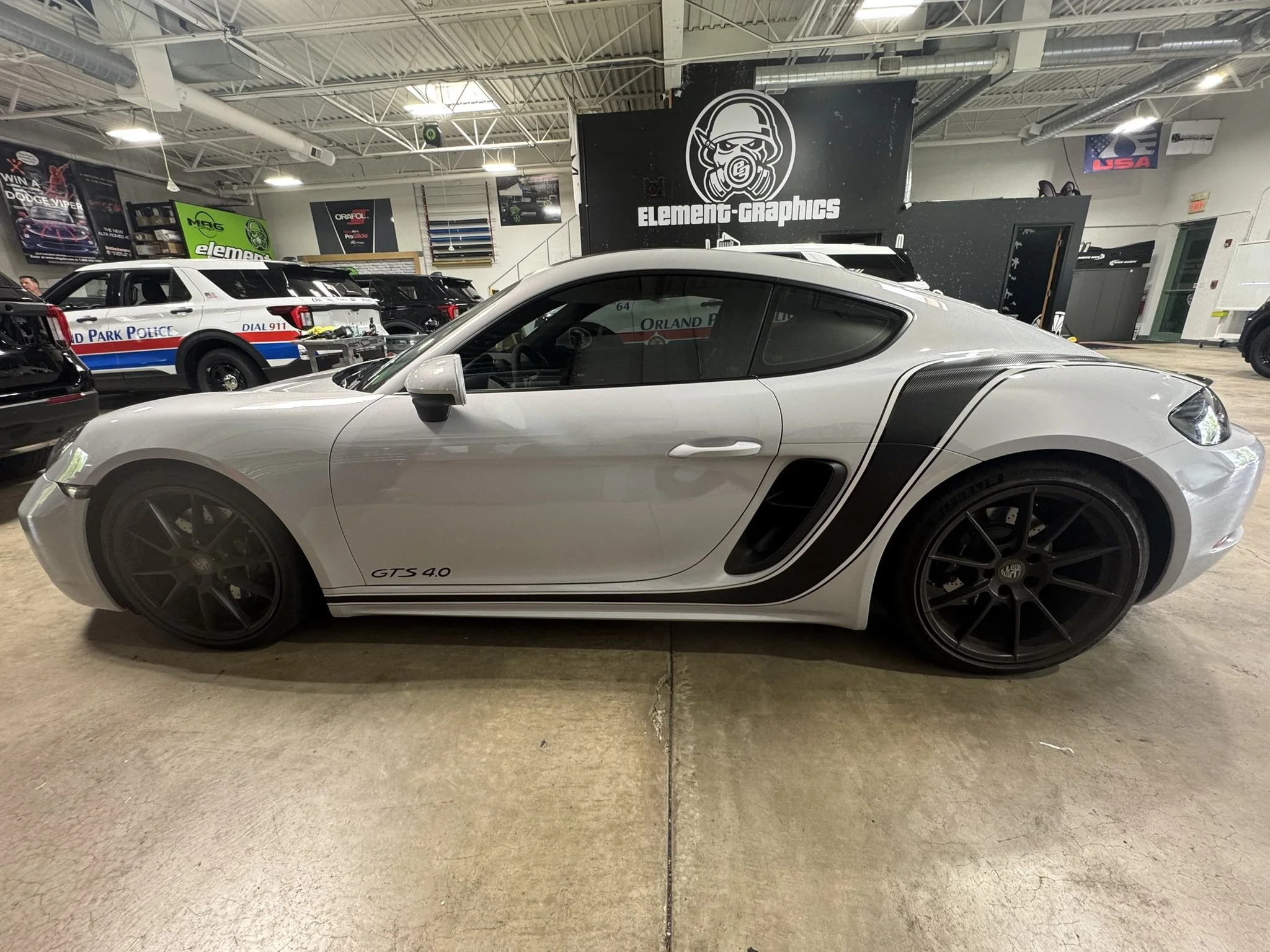 A silver sports car, a Porsche 718 GTS 4.0, parked in an indoor parking lot with other vehicles and promotional banners in the background.