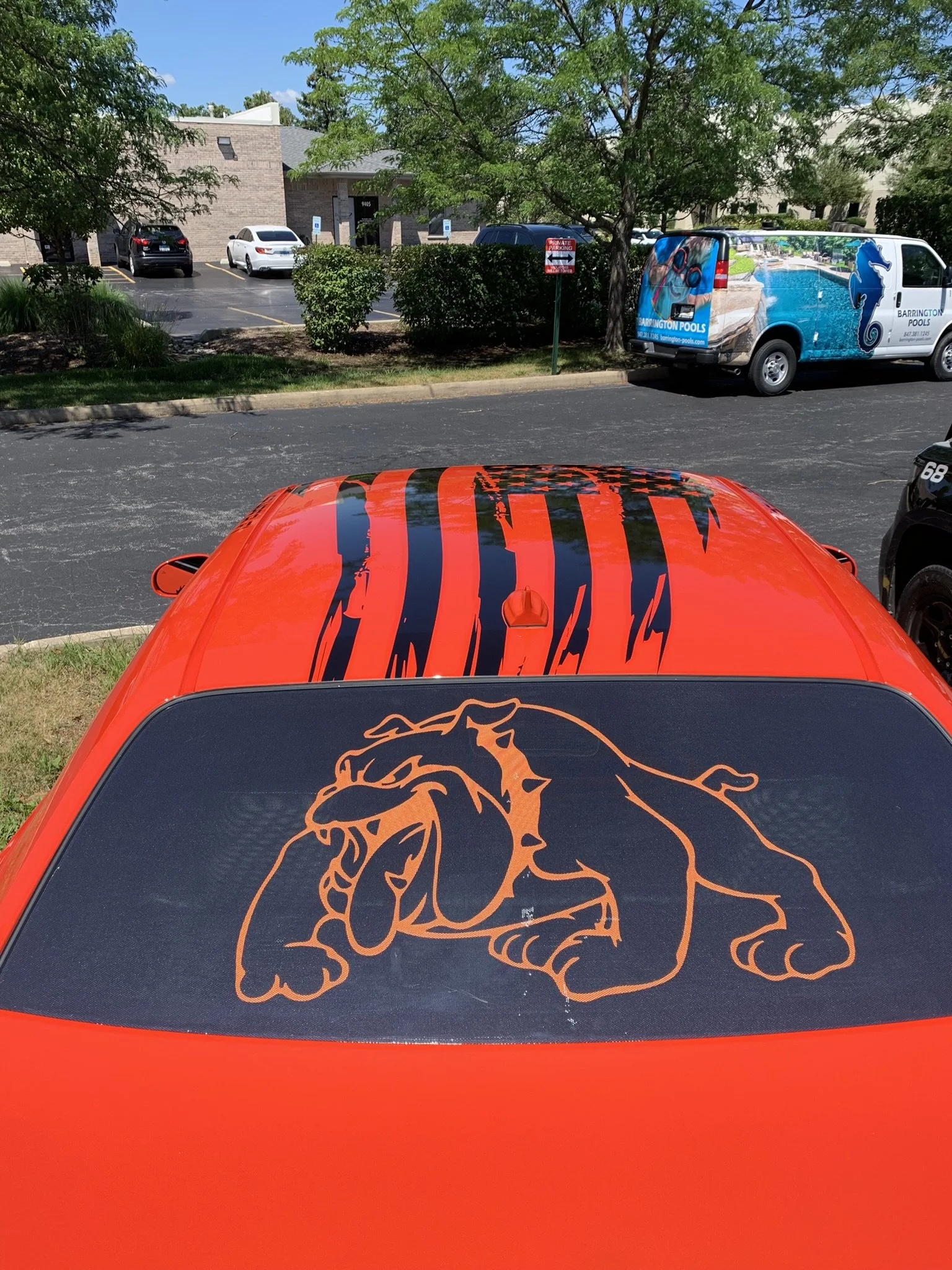 The image shows the back of a red car with a black and orange lion logo on the rear window. In the background, there is a parking lot with several parked cars, a building, trees, and a white van with a cheetah graphic and the text 'Barrington Pools' 
