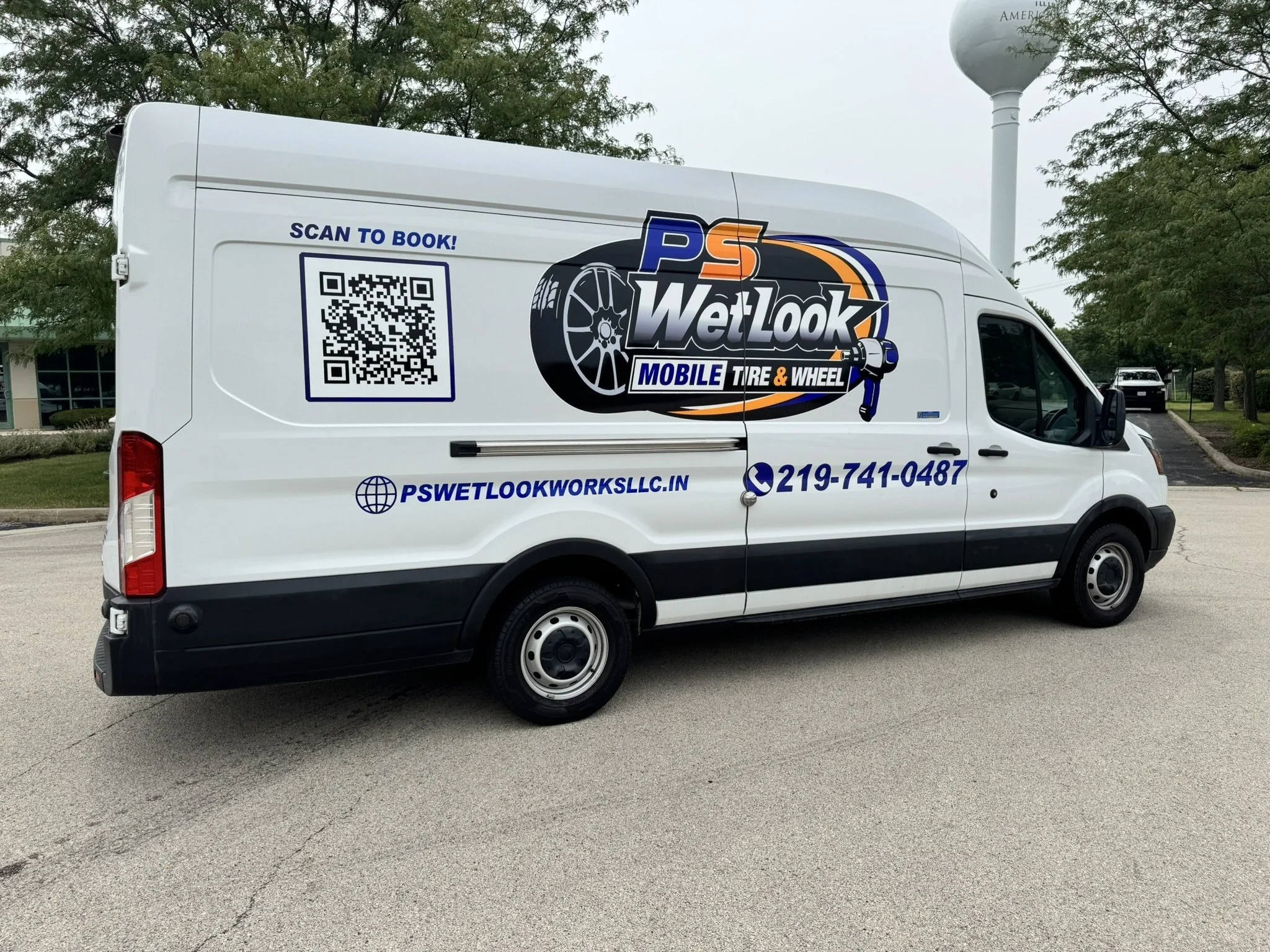 White mobile tire and wheel service van with company logo, QR code, website, and phone number parked outdoors near trees and water tower.