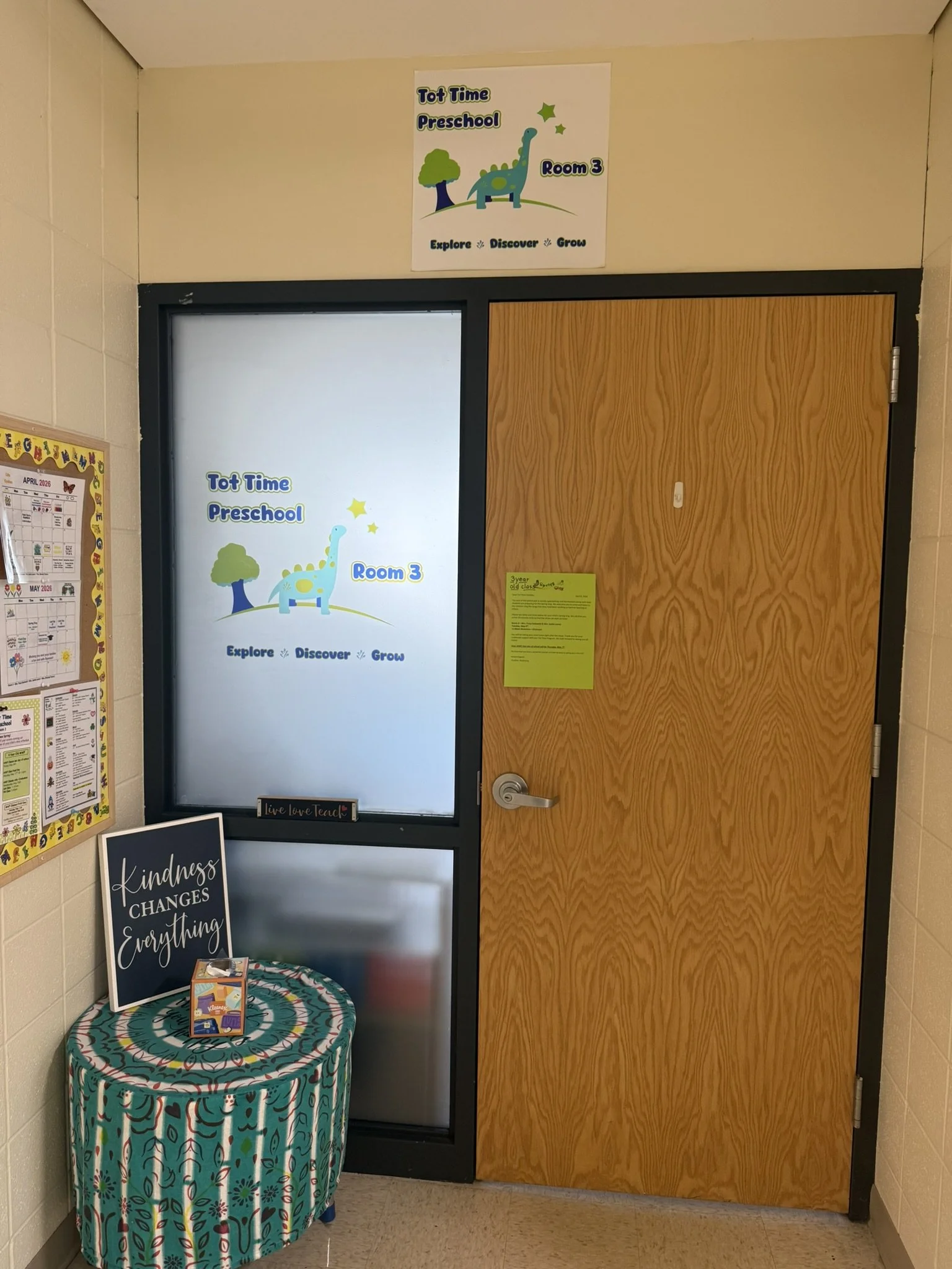 A school hallway with a poster above a door that says "Tot Time Preschool Room 3" and features a cartoon dinosaur and tree. To the left, a bulletin board with schedules or notices. In front, a colorful, patterned table with a sign that reads "Kindnes