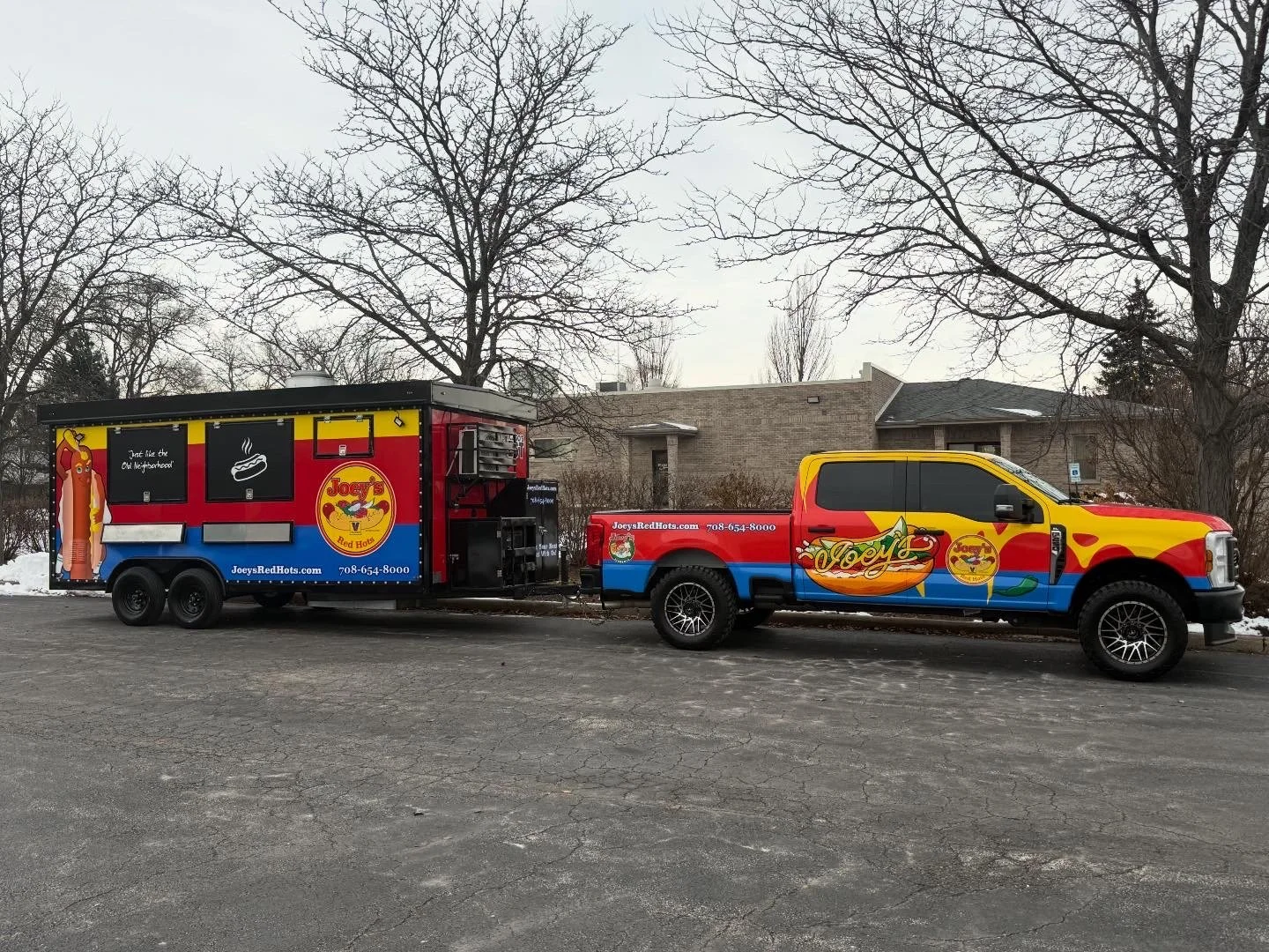 Red and yellow catering truck with chili pepper design parked next to a matching pickup truck, both branded with Joey's logo and website, in a parking lot with trees and houses in the background.