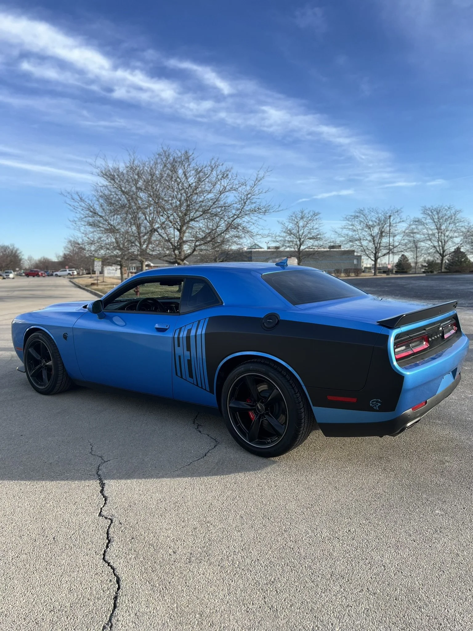 A blue Dodge Challenger car parked in a lot with leafless trees and a clear sky in the background.