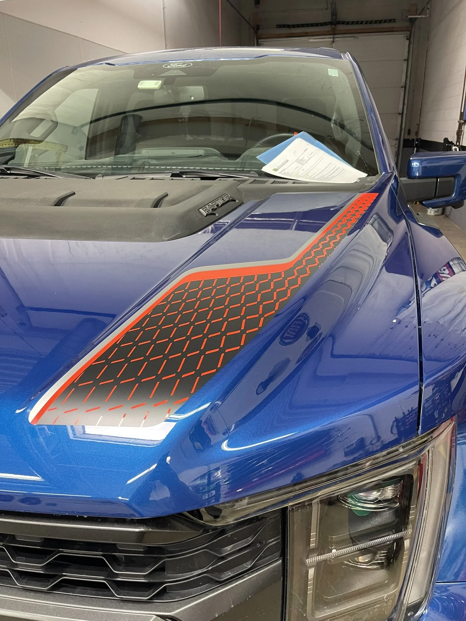 A blue Ford F-150 Raptor pickup truck parked inside a garage, with a sheet of paper on the dashboard visible through the windshield.