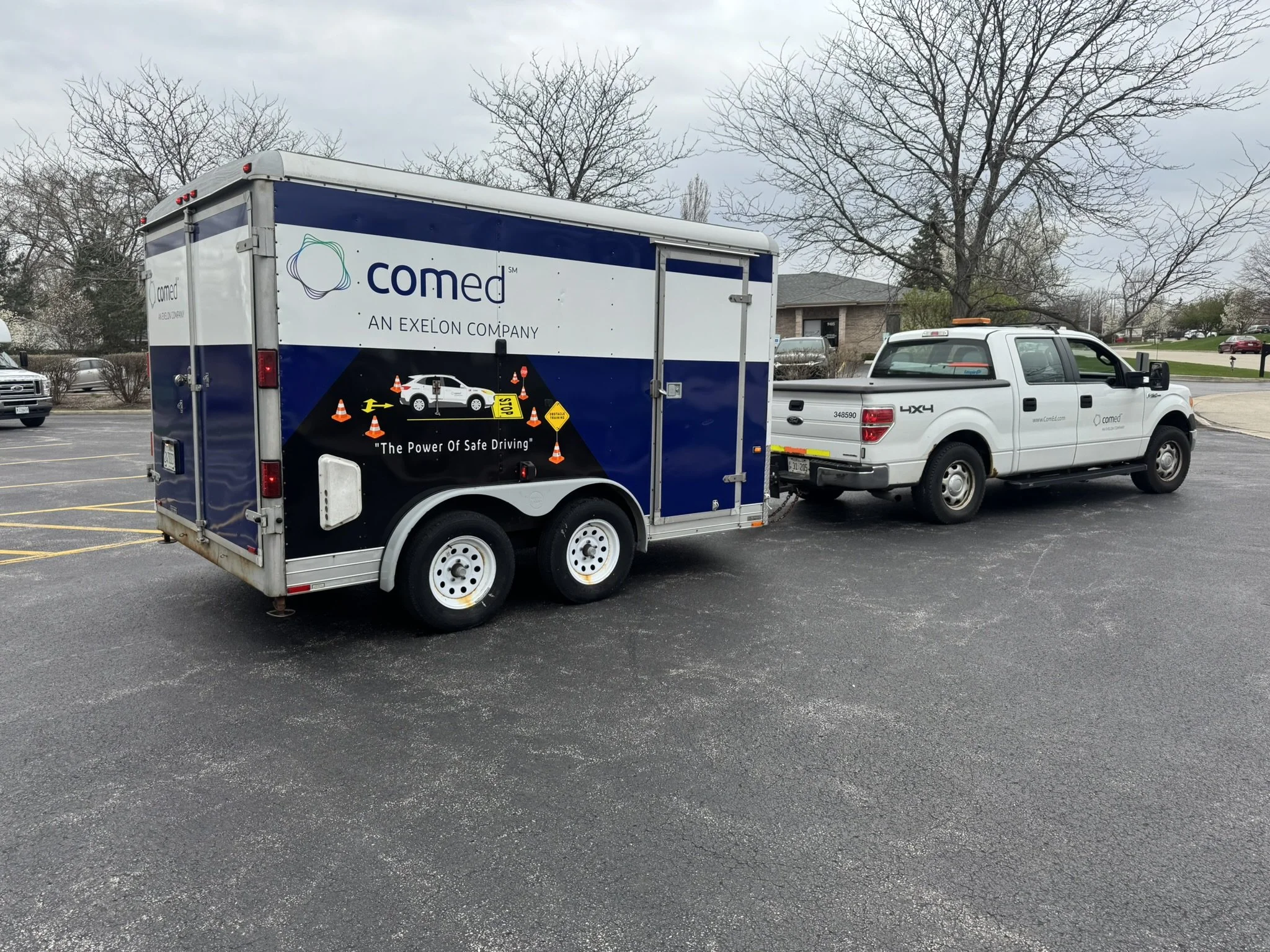 A mobile health trailer with the Comed logo and slogan 'The Power of Safe Driving' parked in a parking lot. There is a white pickup truck attached to the trailer, and several other vehicles and trees in the background.