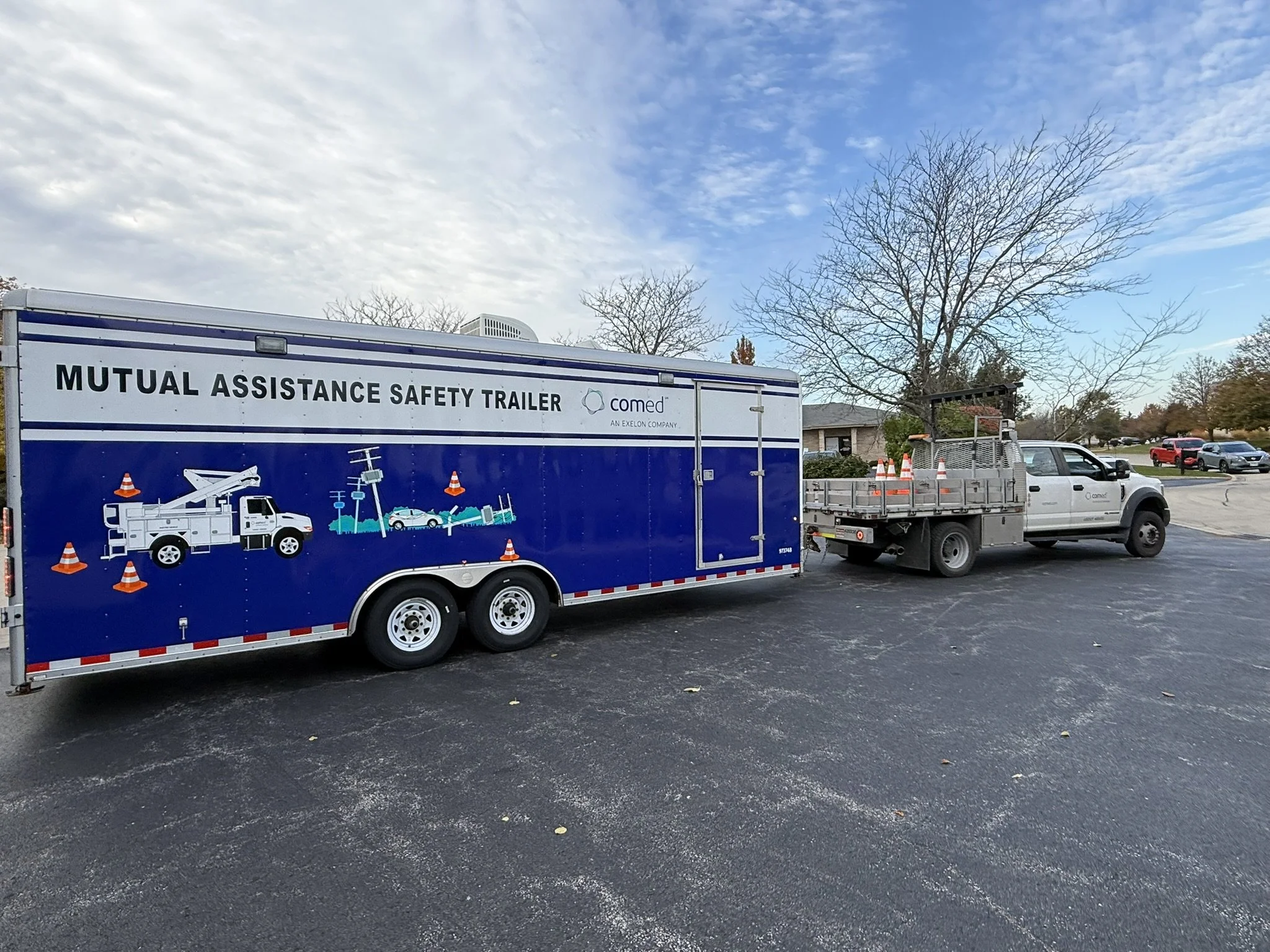 A blue trailer labeled 'Mutual Assistance Safety Trailer' parked on an asphalt lot with orange traffic cones, a white pickup truck, and other construction equipment nearby. Leafless trees and a cloudy sky are visible in the background.