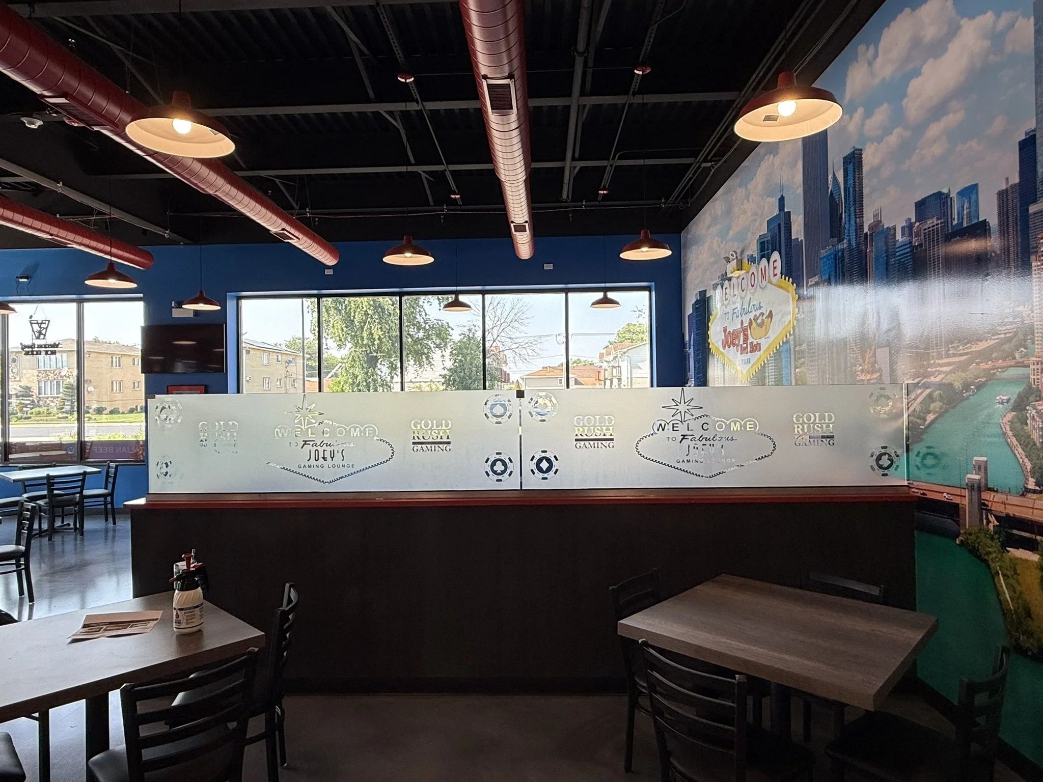 Interior of a gaming lounge with tables and chairs, large window with a city view, and cityscape mural on the wall.