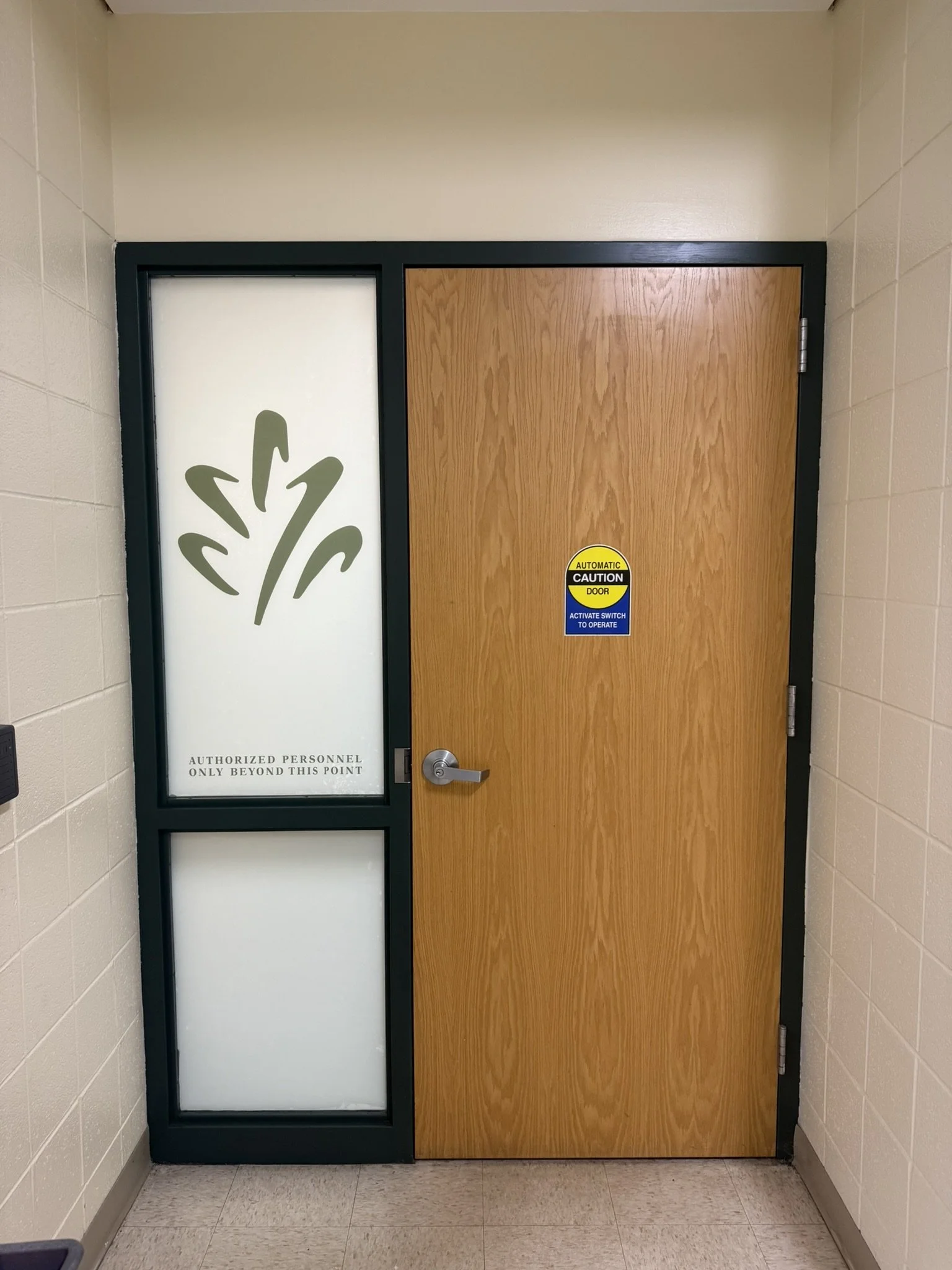 A double door with a wood grain finish on the right and a frosted glass panel on the left, set in a black frame. The frosted glass panel has a decorative plant design and the words "AUTHORIZED PERSONNEL ONLY BEYOND THIS POINT". There is a yellow and 