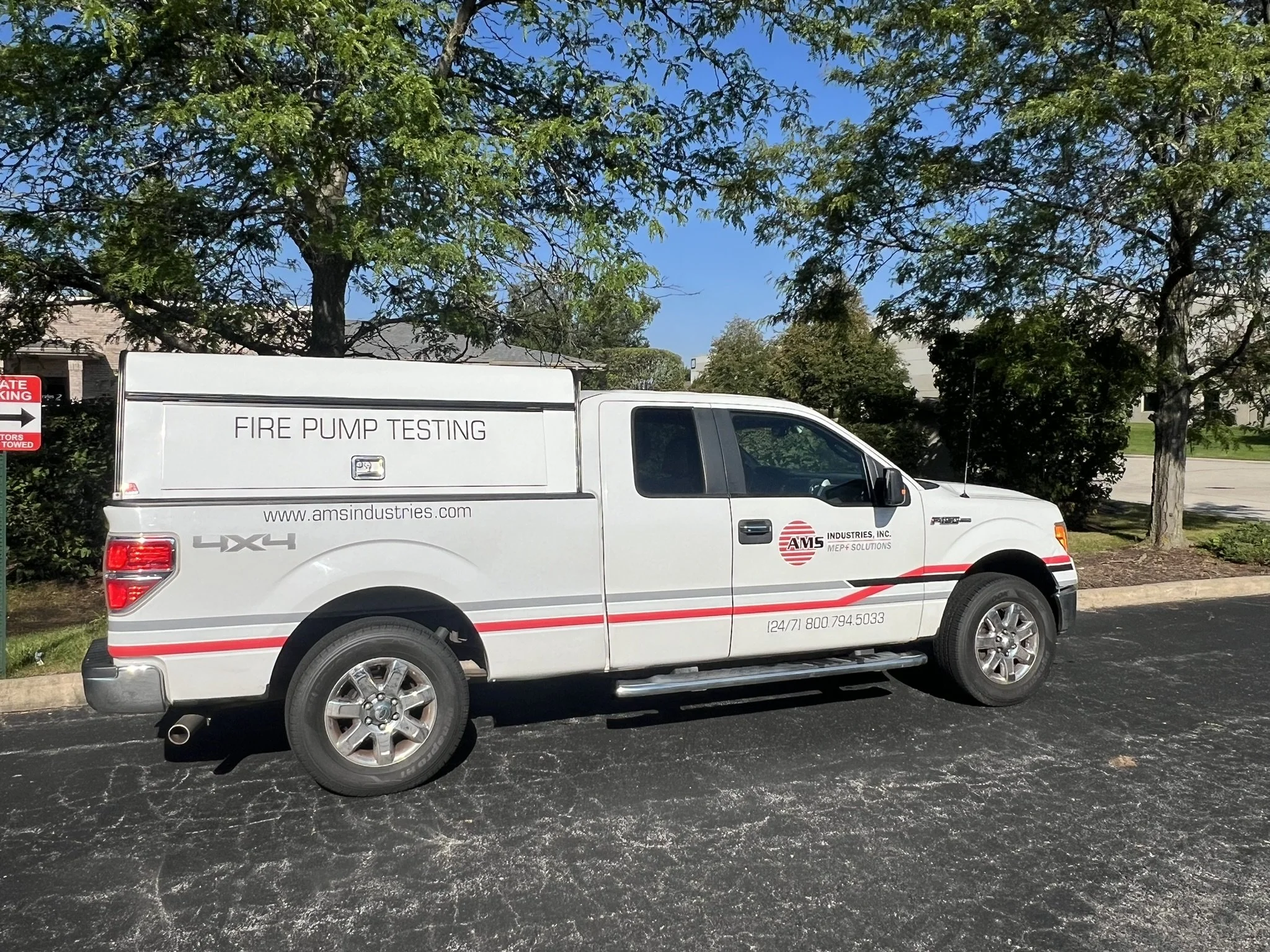 White utility vehicle with 'FIRE PUMP TESTING' written on the side, parked on asphalt with trees and houses in the background.