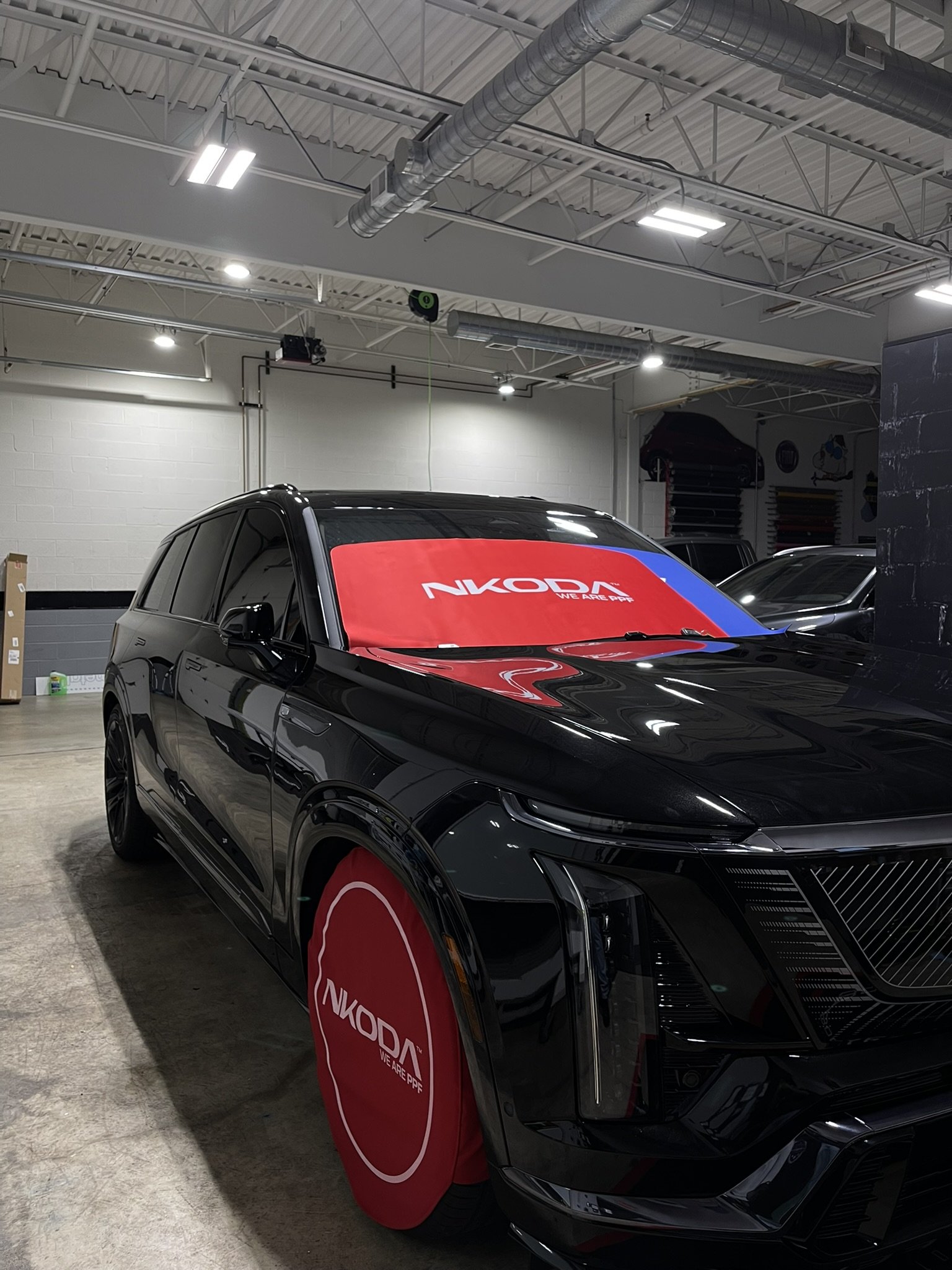 A black electric car parked indoors at a dealership or showroom, with a red and blue windshield cover displaying the NikoD logo and slogan.