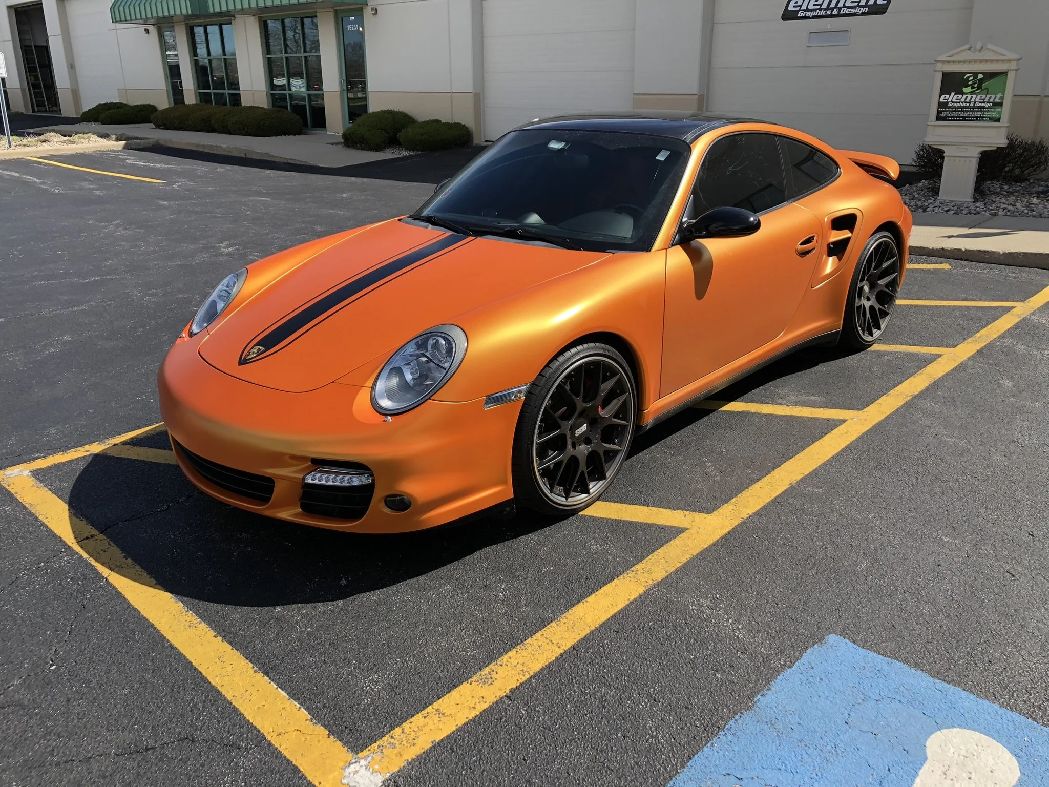 Orange sports car parked in a parking lot with black racing stripes on the hood, near a building with bushes and signs.