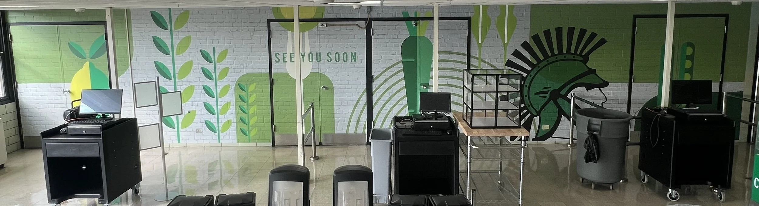 Empty airport check-in counter area with a colorful mural of green plants, a large black warrior helmet mural, and a sign that says 'SEE YOU SOON' on the wall.
