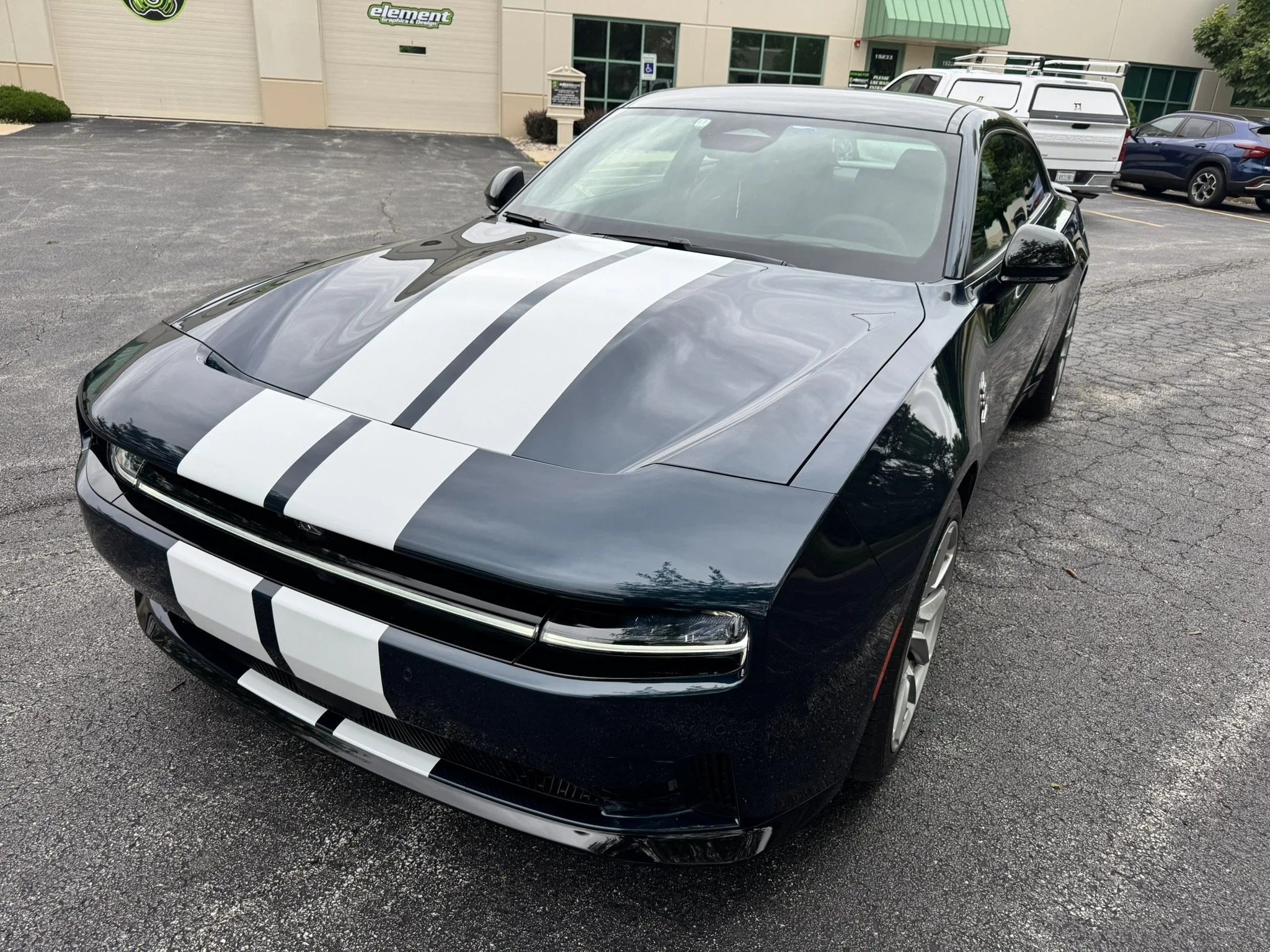 A black sports car with white racing stripes parked in a parking lot outside a building with a green awning and an 'element' signage. Other vehicles are parked nearby.