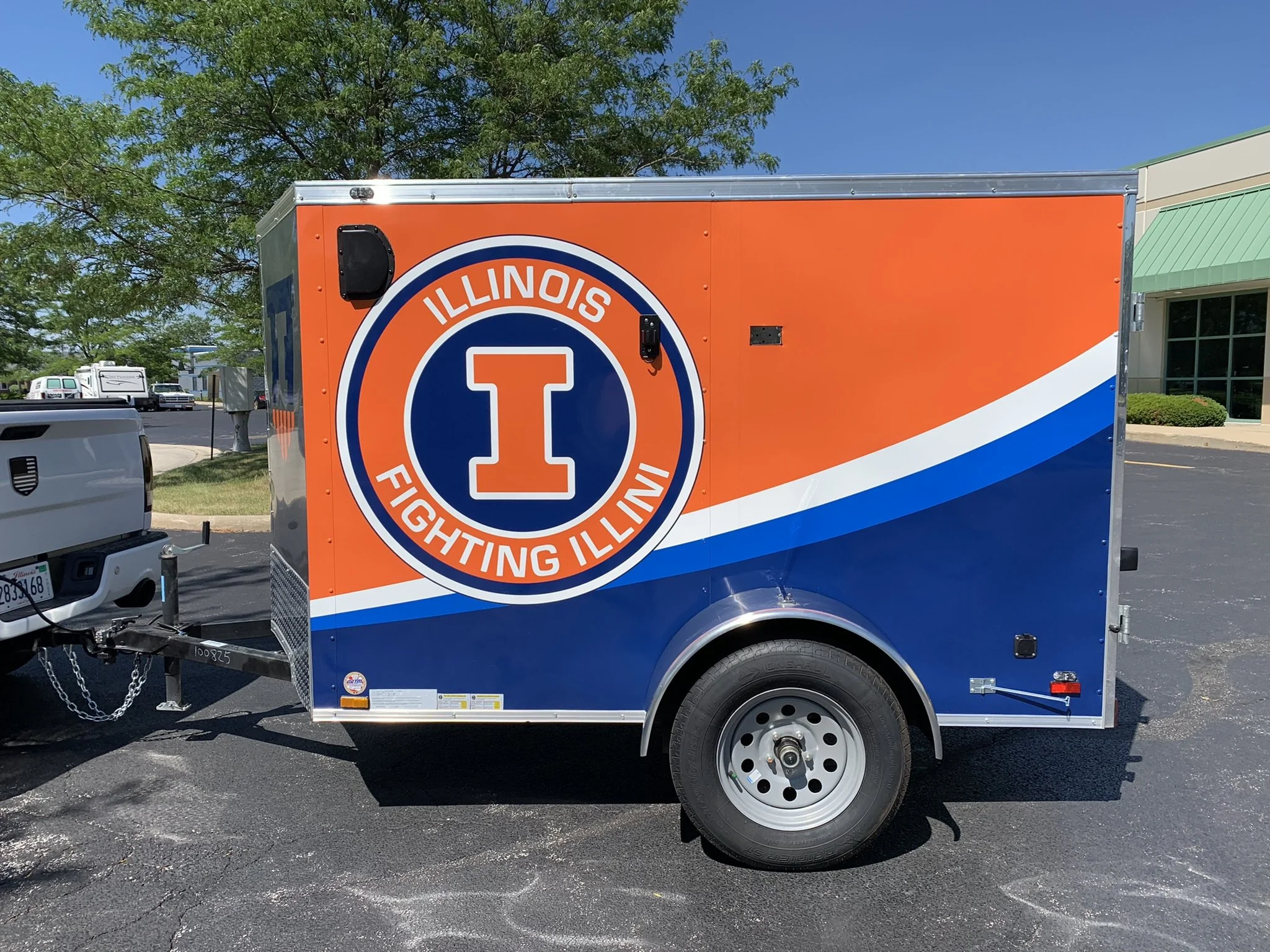 A trailer with the Illinois Fighting Illini logo and colors parked on asphalt, with trees and a building in the background.