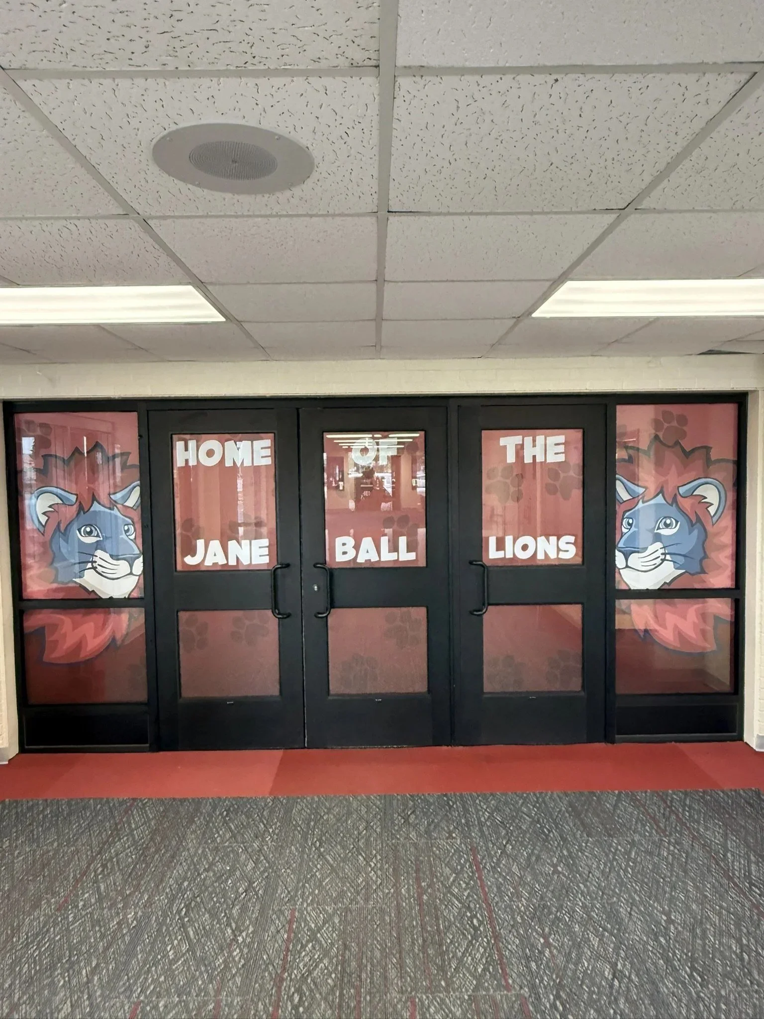 Double door with posters of a blue lion cartoon on each side, and words "Home of the Jane Ball Lions" on the posters, in a school hallway with a speckled gray floor.
