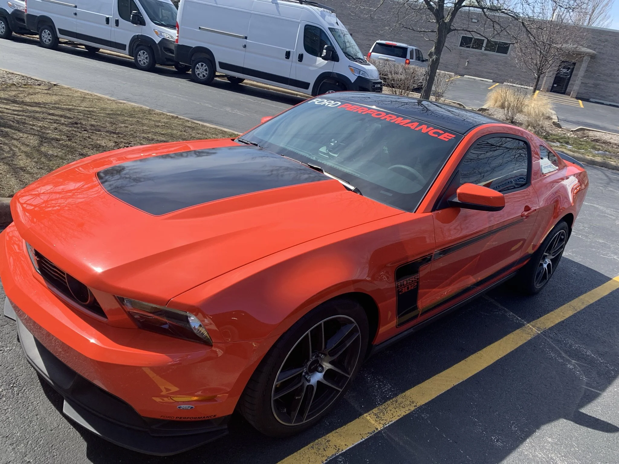 Close-up view of a red Ford Mustang with black racing stripes, parked in a parking lot. The windshield has a banner that reads 'Ford Performance' in red and white letters.