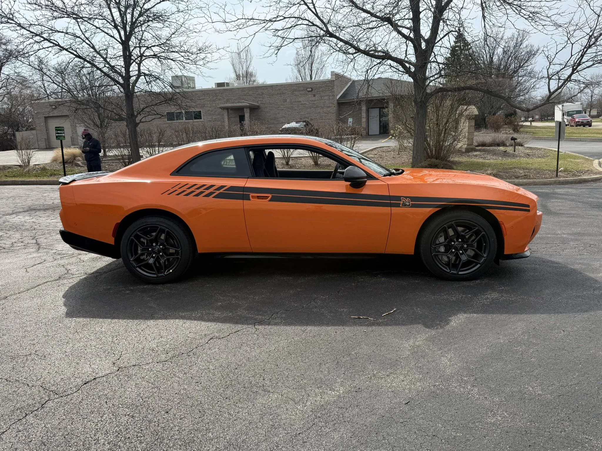 A bright orange sports car with black stripes is parked on a street in front of a building with leafless trees and a person wearing a winter hat and coat.