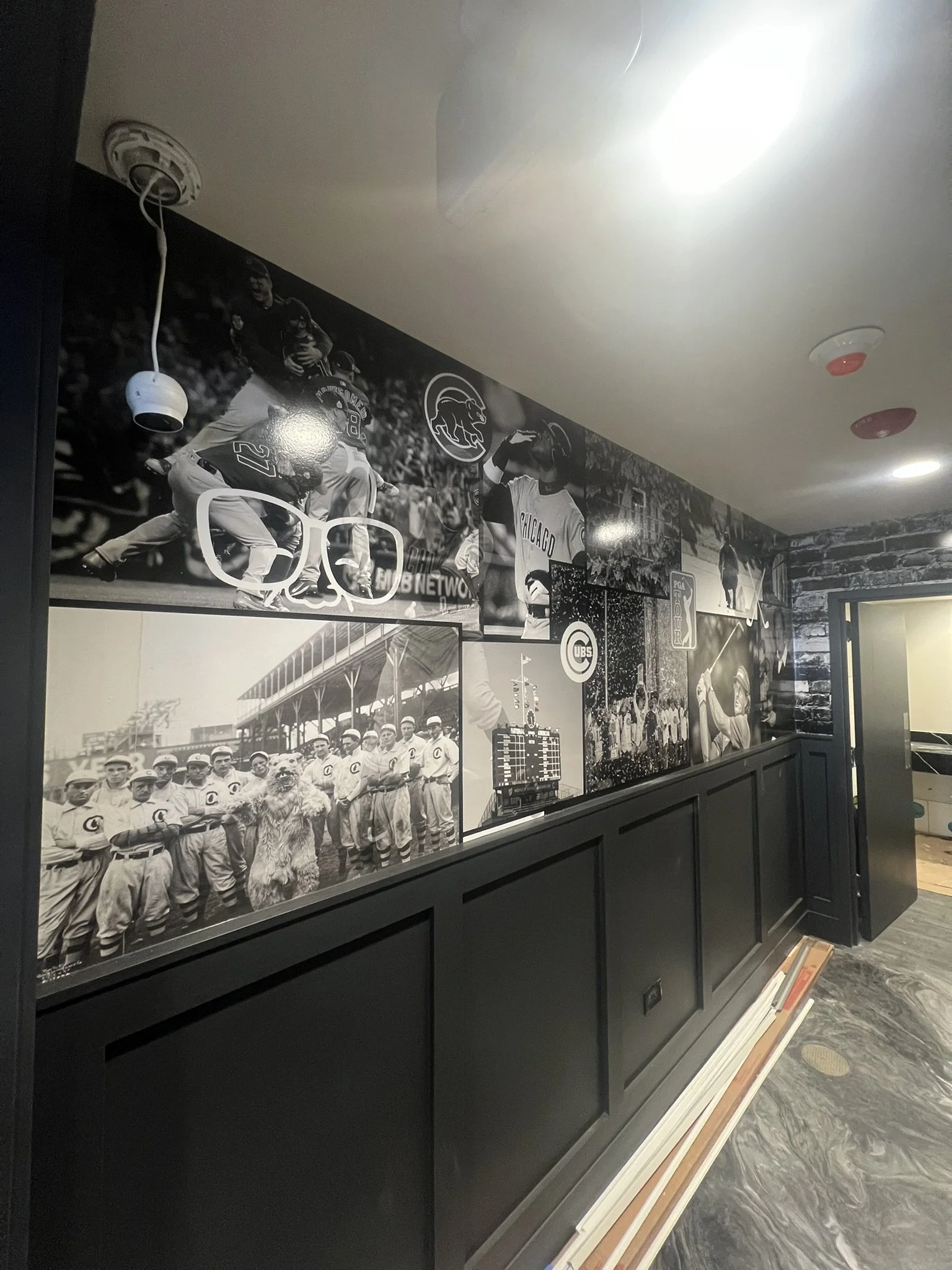 Black and white collage of vintage baseball photos, featuring players in uniforms, a team photo, and various action shots, displayed on a wall with a black wainscot.