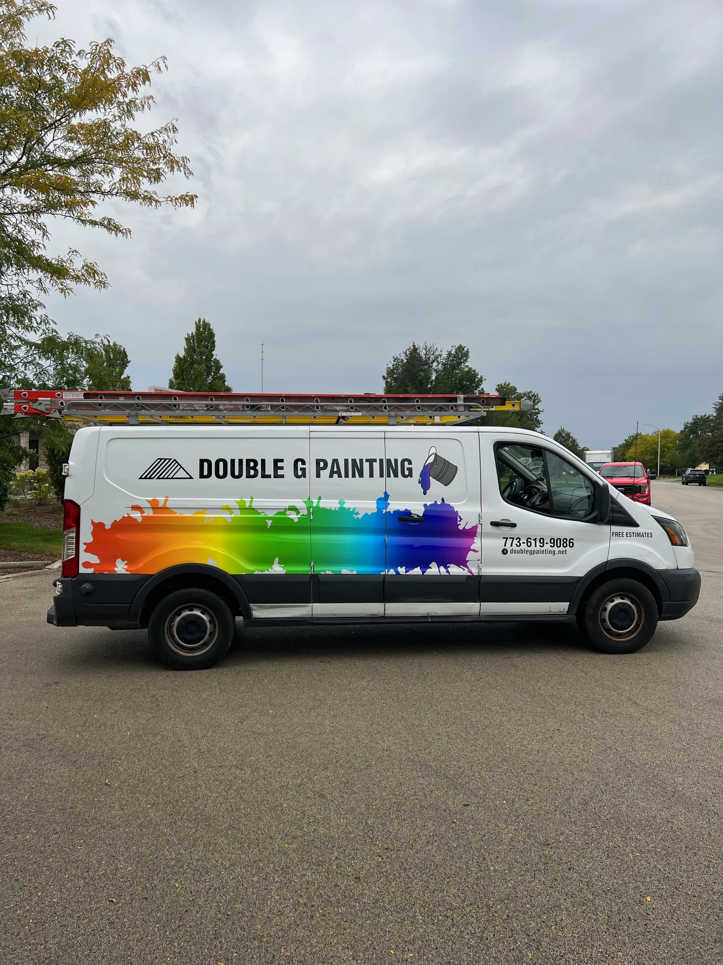 A white service van with colorful rainbow paint splashes and the text 'Double G Painting' parked on a paved lot with trees and cars in the background.
