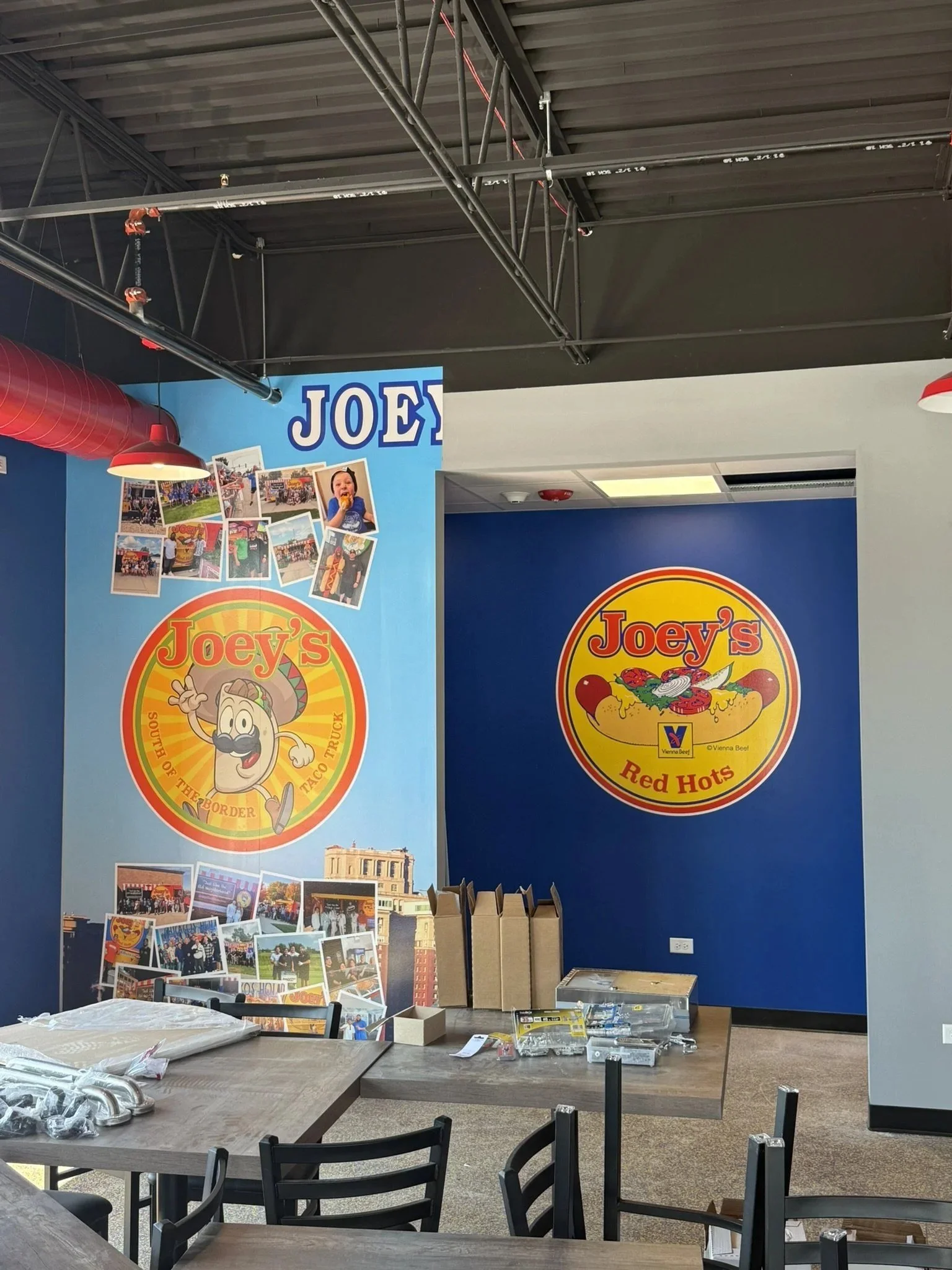 Interior of a restaurant or cafe with tables and chairs, decorated with Joey's branding, including colorful posters and banners on the wall featuring a cartoon mascot and photos of events.