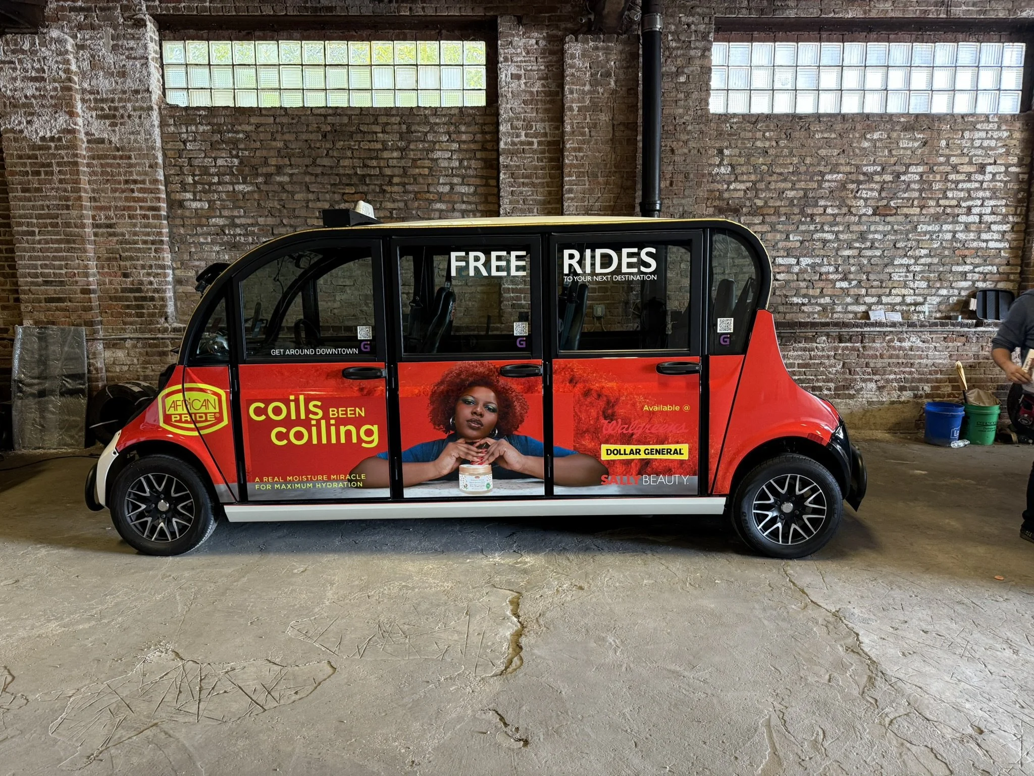 A red electric vehicle with advertising on its side, promoting free rides and hair care products, parked inside a building with exposed brick walls, high windows, and a concrete floor.
