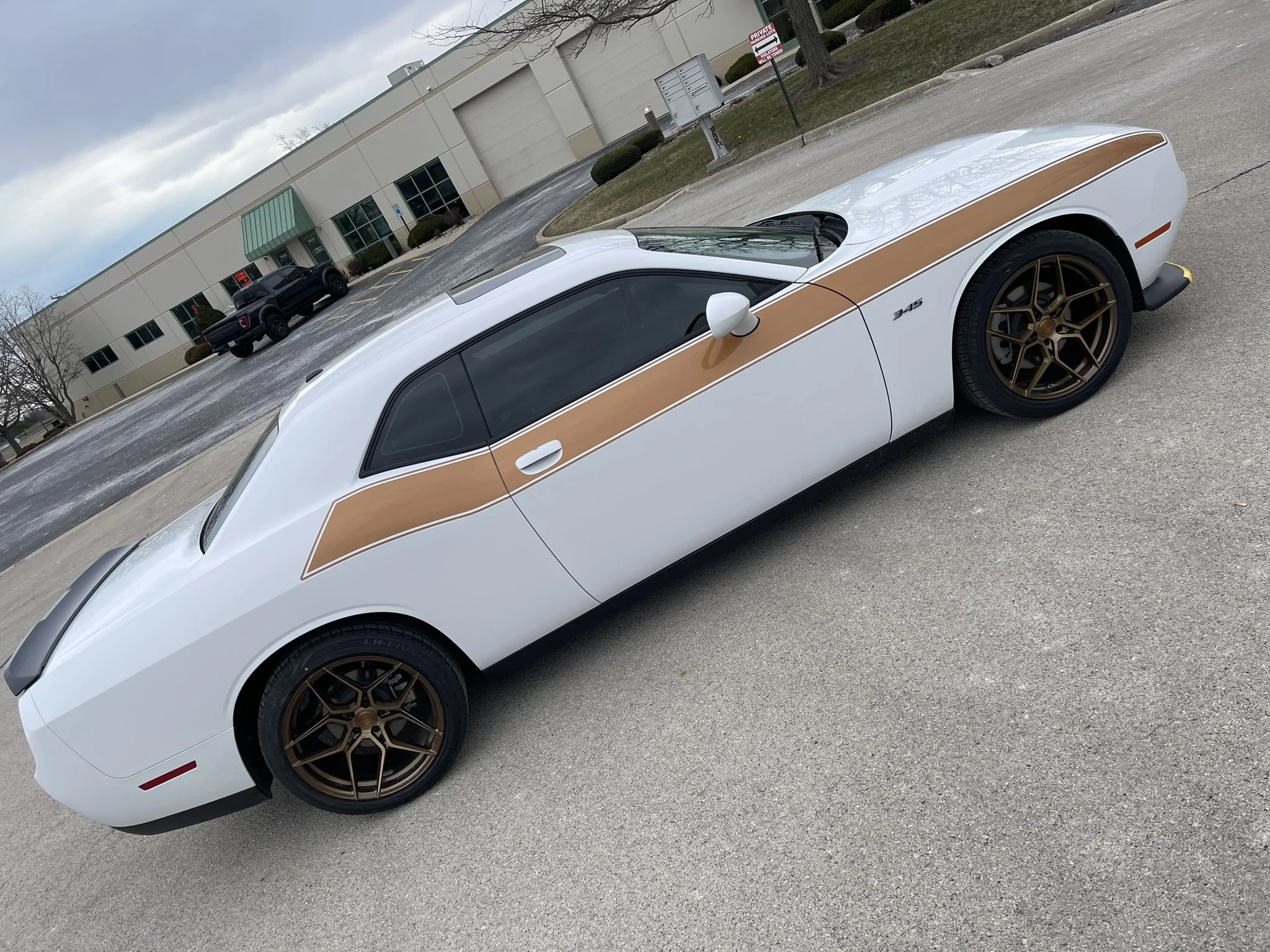 A white sports car with gold wheels and a tan stripe along the side parked in a parking lot outside a commercial building on a cloudy day.