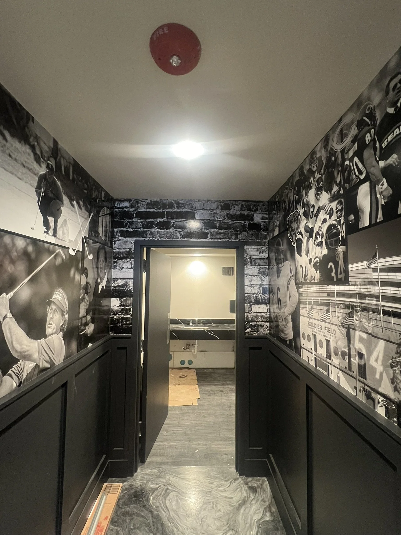 A hallway with black paneling and black-and-white sports-themed murals on the walls, leading to a room with a sink and a mirror. The ceiling has a red fire alarm, and the floor is marbled gray.