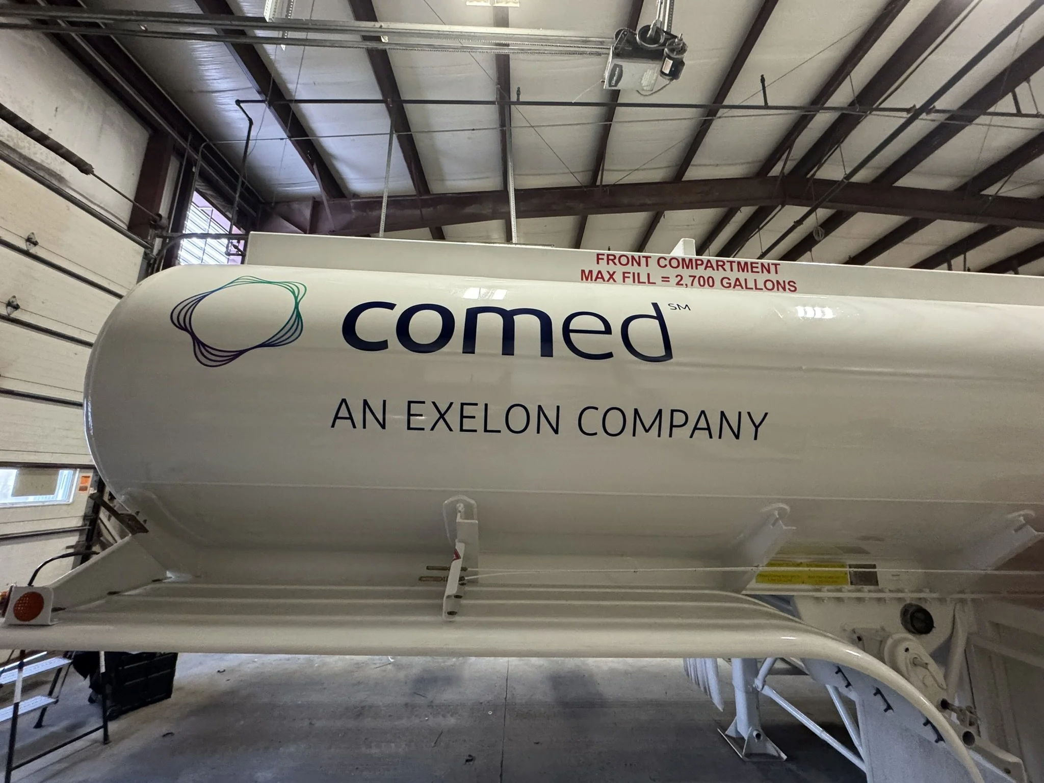 The image shows the underside of a white boat inside a warehouse. The boat has the logo "comed" and the text "AN EXELON COMPANY". There is a sign on the boat indicating the front compartment's maximum fill capacity of 2,700 gallons. The warehouse has