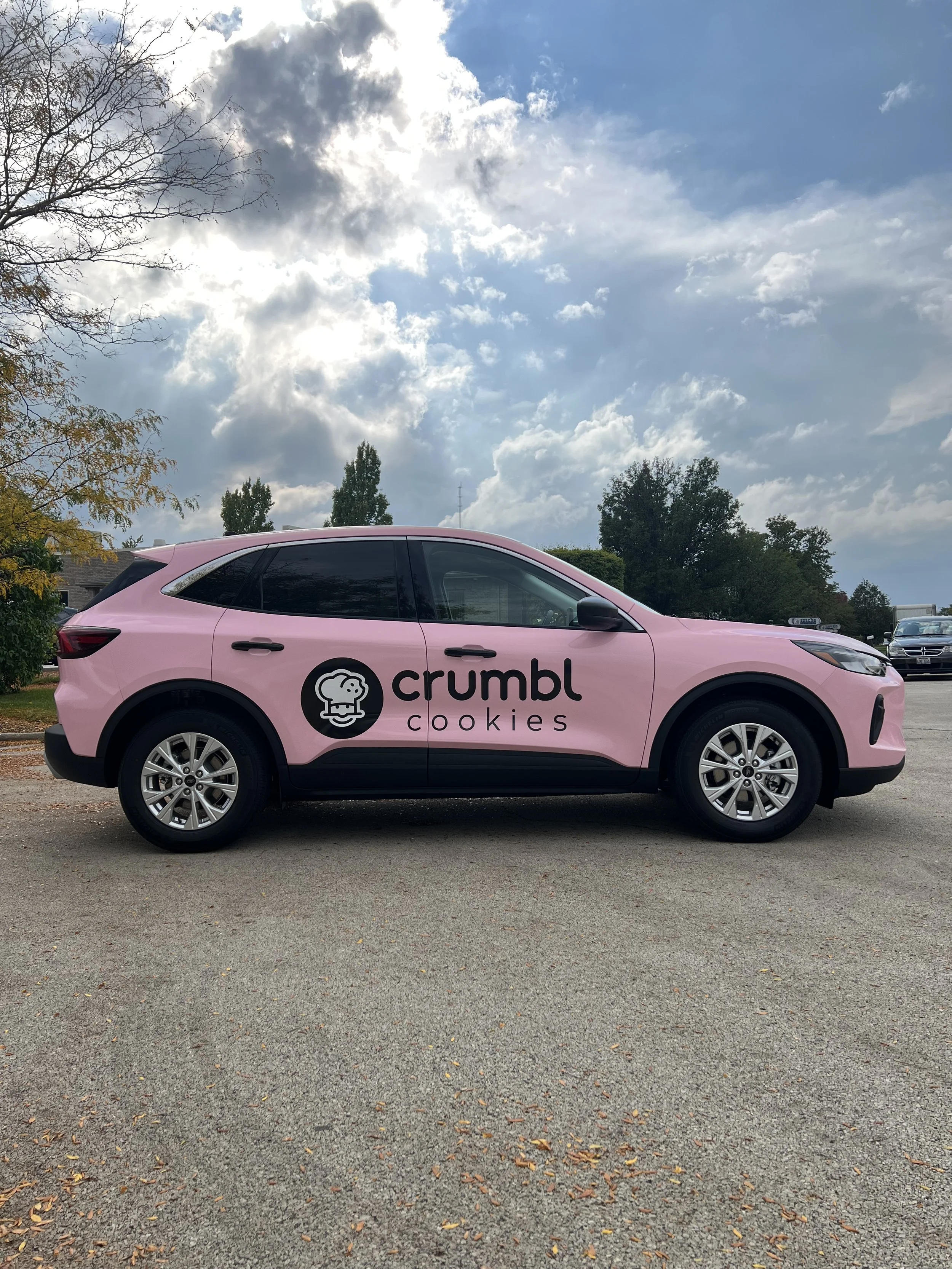 A pink SUV with "crumb cookies" logo on the door, parked on a paved surface with fallen leaves. The background shows a partly cloudy sky and some trees.
