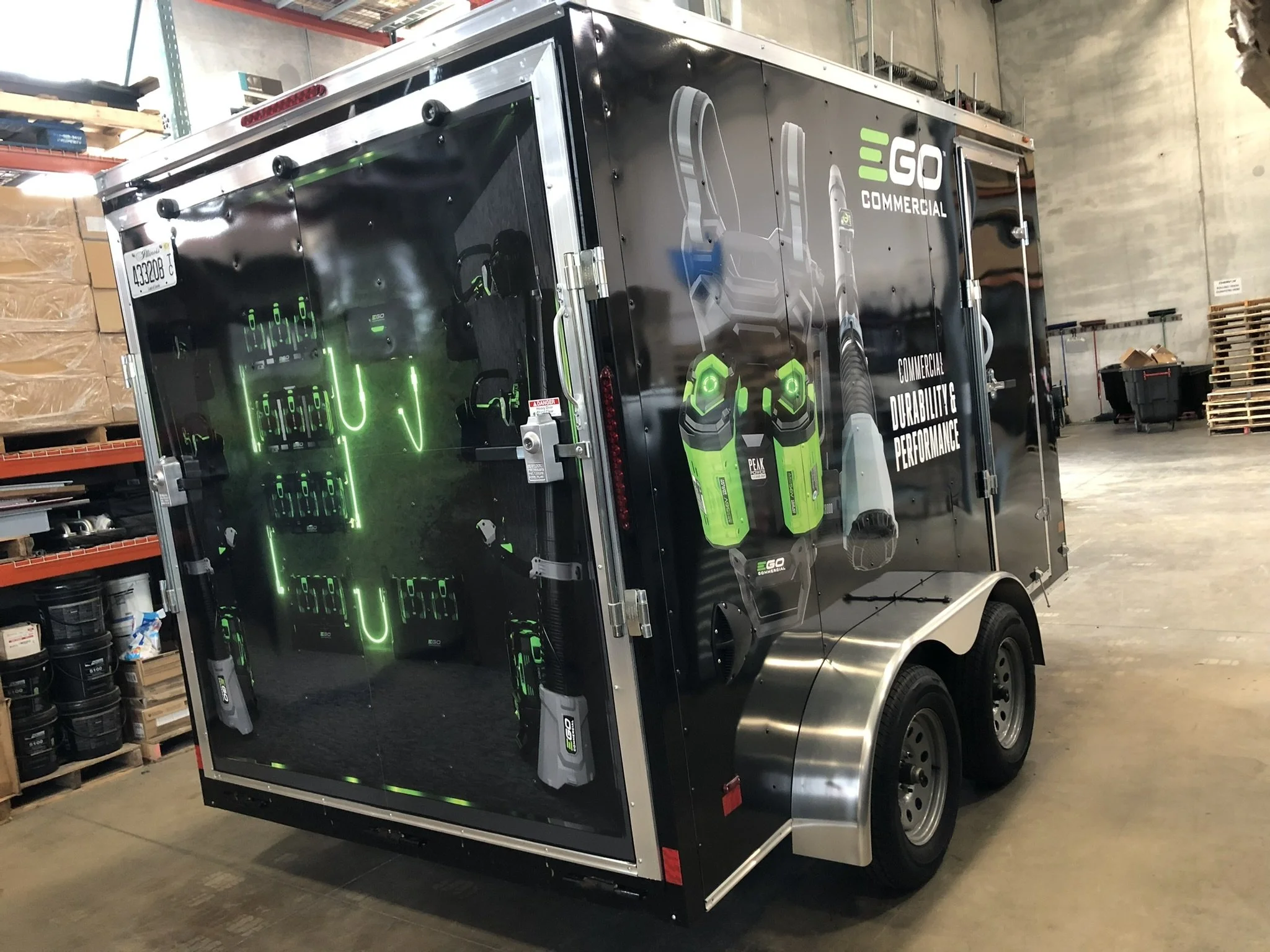 A black mobile vending trailer with a graphic ad on the side displaying green and gray battery images and the text 'GO Commercial', 'Durability & Performance'. The trailer is in a warehouse, surrounded by shelves and various items.