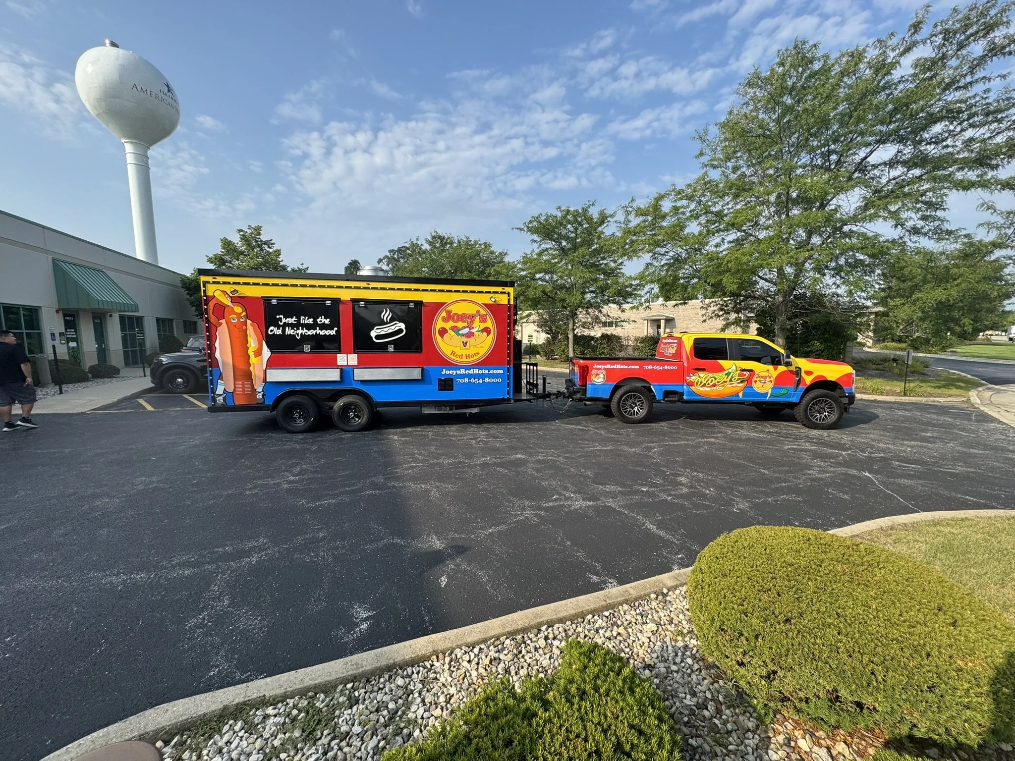 A parking lot with a water tower in the background, two colorful food trucks parked side by side, trees, and bushes in the surroundings.