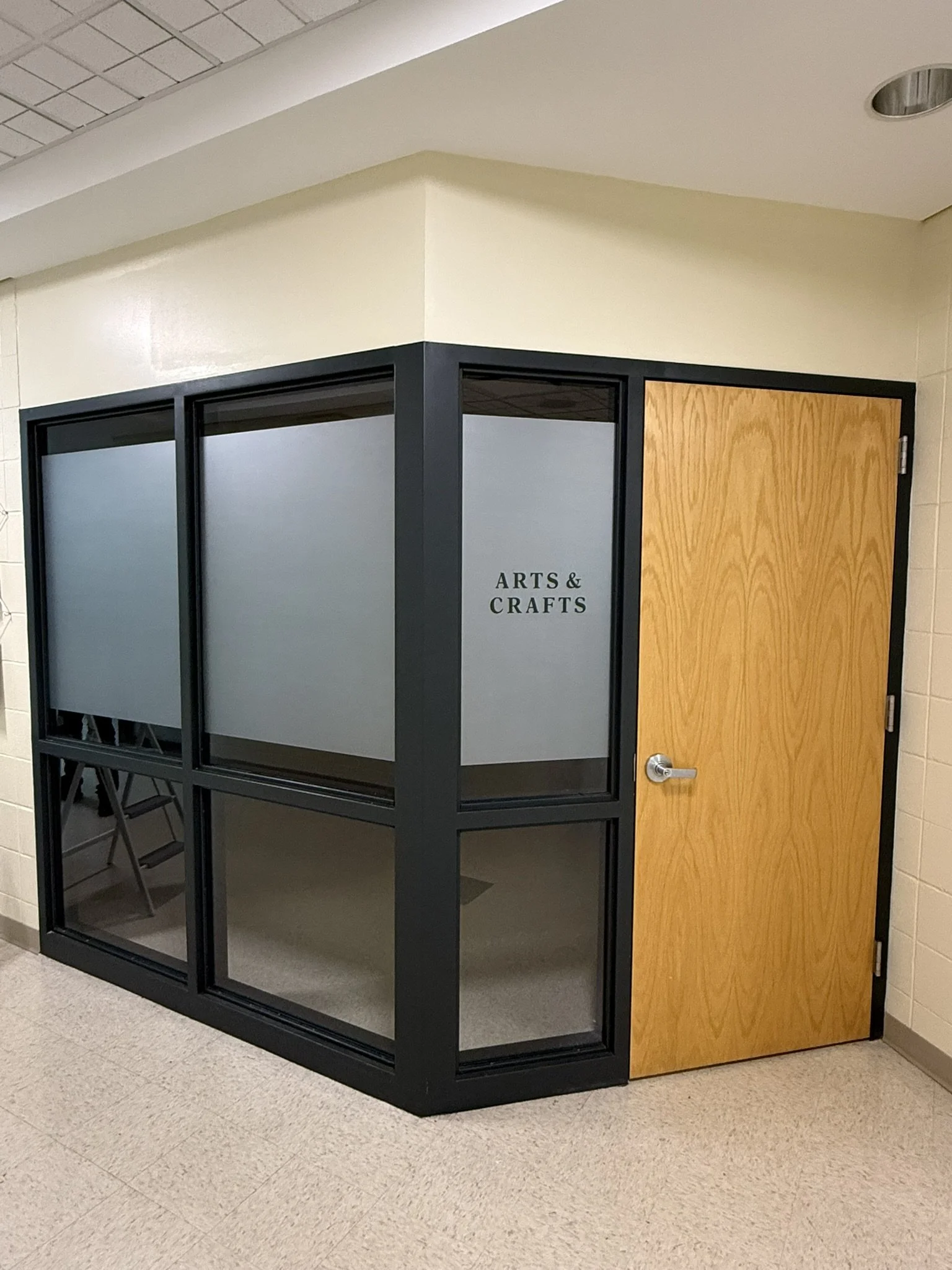 Indoor area with a black-framed glass partition labeled 'Arts & Crafts' and a wooden door beside it.