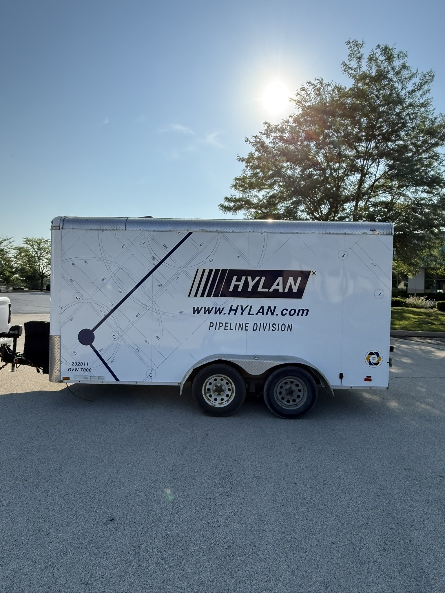 A white trailer with the Hylan logo and website, parked on a paved surface with a tree and blue sky in the background.