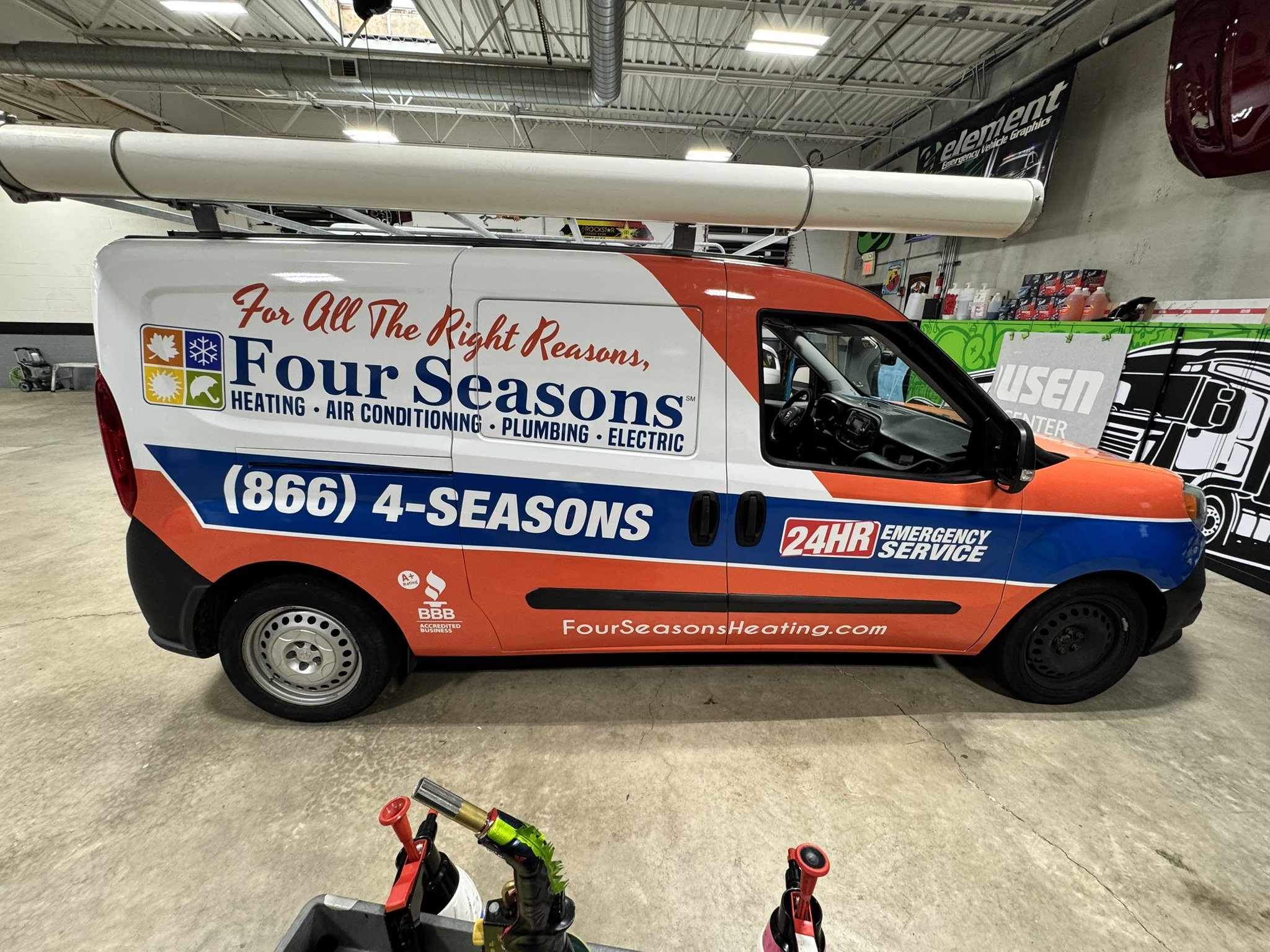 A company van for Four Seasons Heating, Air Conditioning, Plumbing, and Electric parked indoors. The van features branding and contact information with a slogan, inside a warehouse or showroom, with various signs and equipment visible in the backgrou