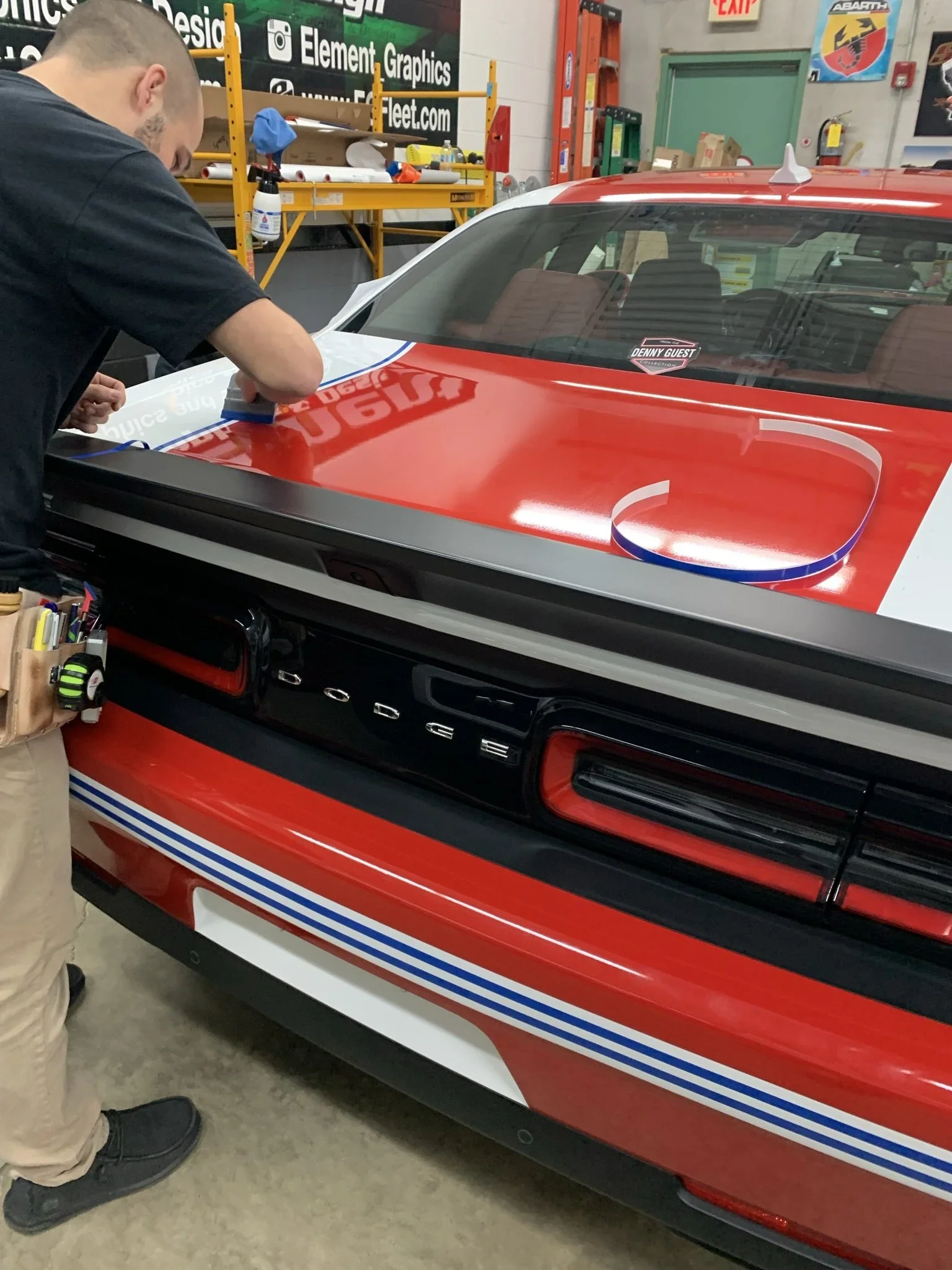 Person applying a decal or sticker to the rear of a red and white sports car inside a workshop or garage.