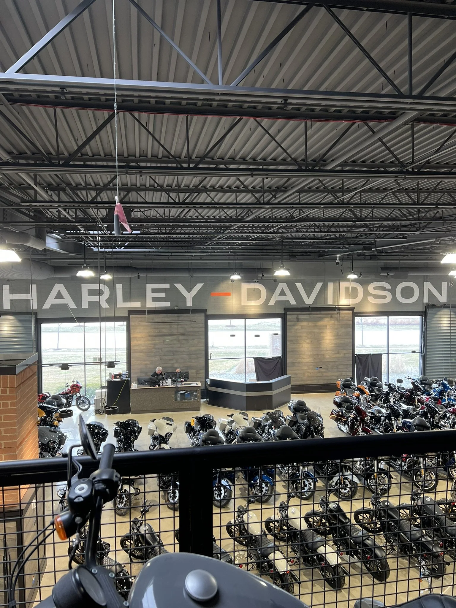 Interior of a Harley-Davidson motorcycle showroom with many motorcycles on display, a counter with staff, large windows, and the Harley-Davidson sign on the wall.