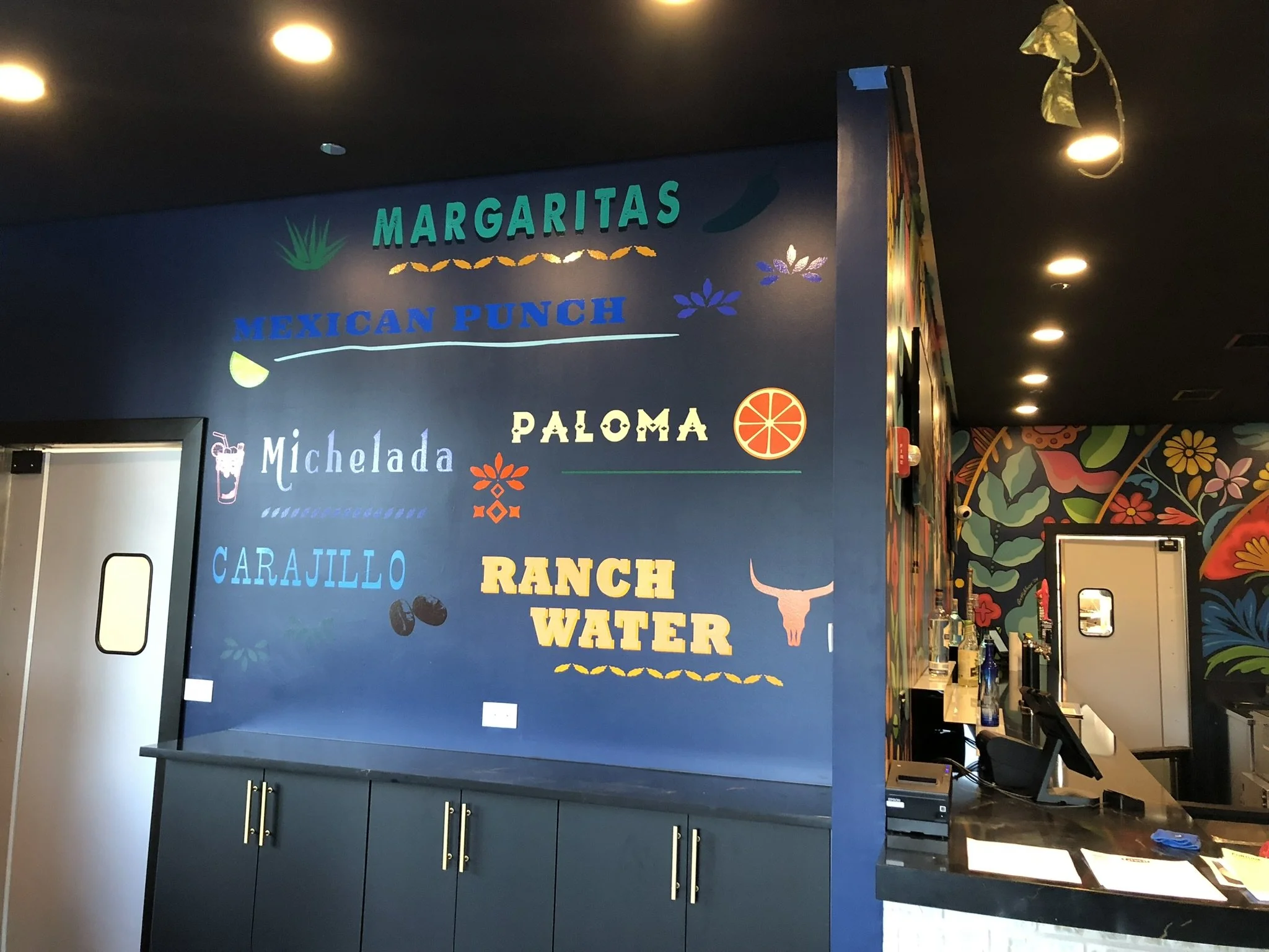 Interior of a restaurant or bar with a blue wall decorated with colorful hand-painted illustrations and text. The text includes words 'MARGARITAS,' 'MEXICAN PUNCH,' 'PALOMA,' 'Michelada,' 'RANCH WATER,' and other menu items. There is a doorway on the