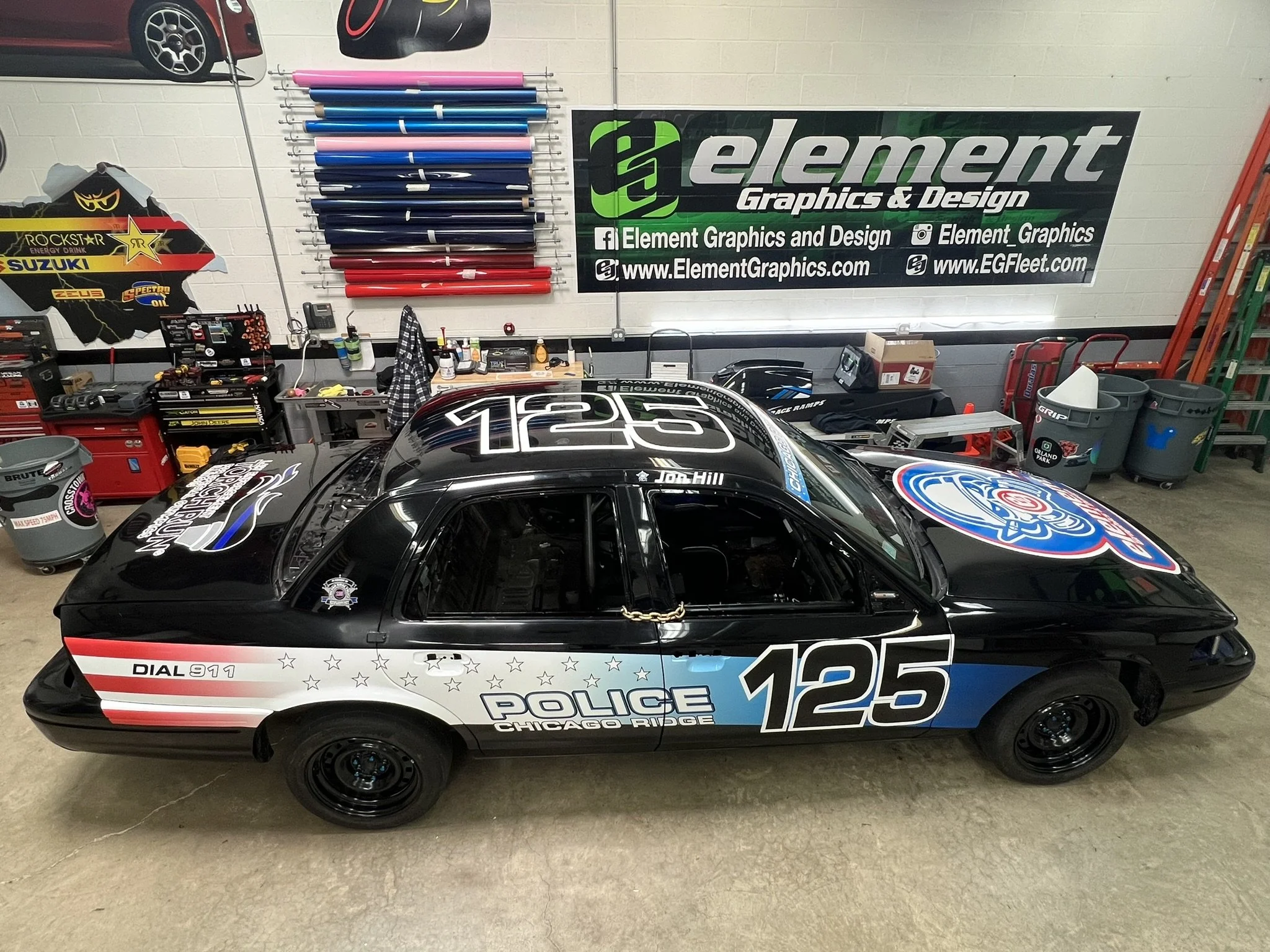 A Chicago police car with a black, white, and blue design, featuring the number 125 and various decals, parked indoors in front of a tool and equipment workspace. The workspace has a large green and black sign for Element Graphics & Design, with rain