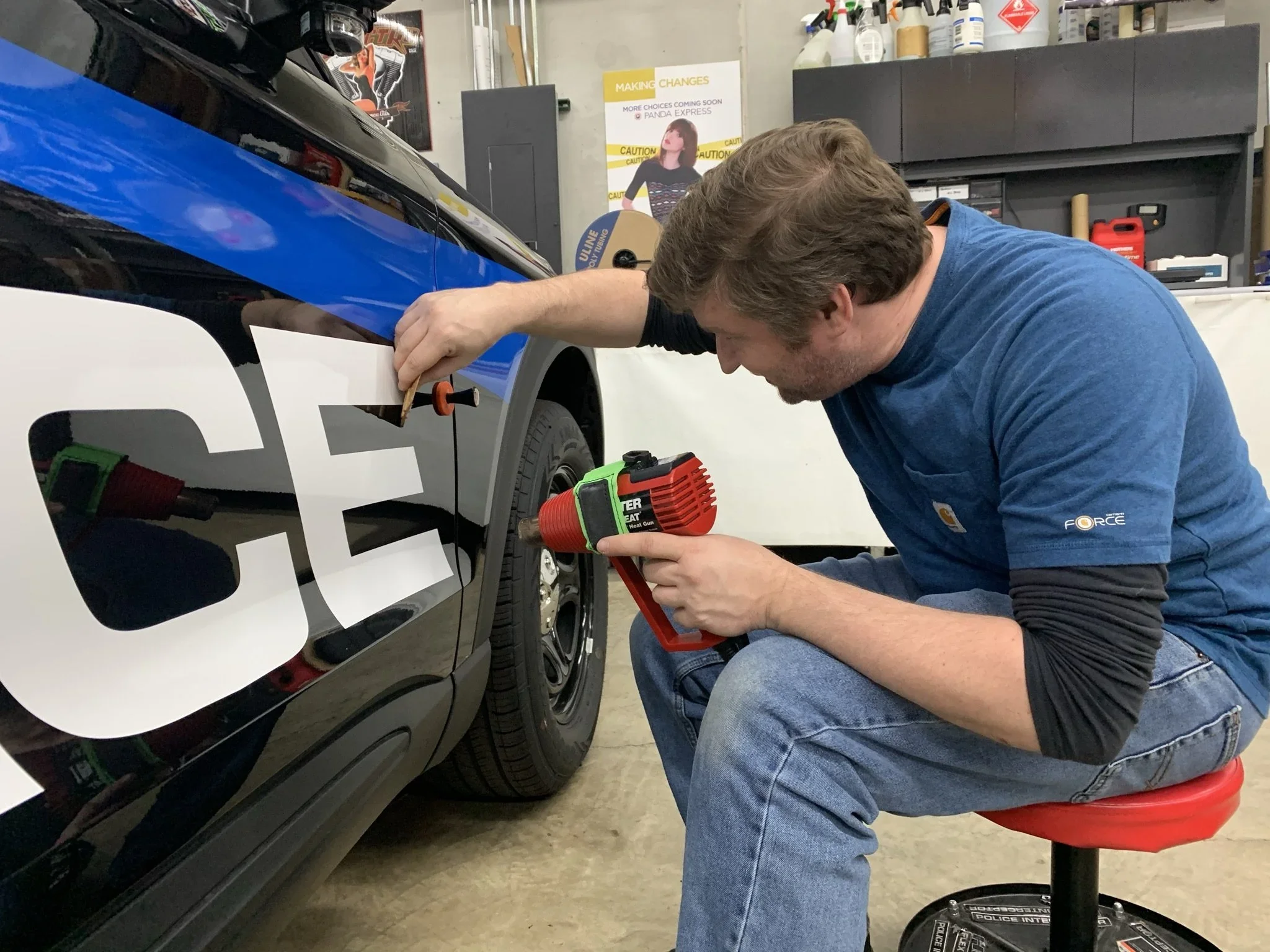 Man kneeling on a red chair, using a heat gun to apply decals to the side of a race car in a garage. The garage has shelves with supplies and a poster in the background.