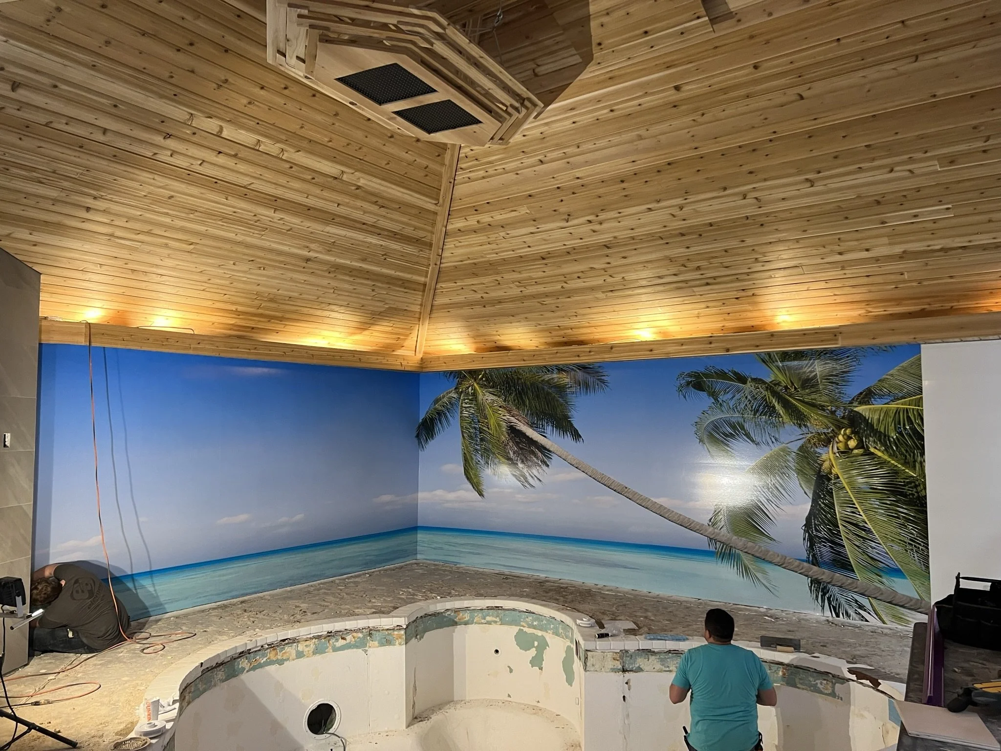 Interior of a room under construction with a tropical themed wall mural of palm trees and the ocean, a wooden ceiling, and two workers installing or repairing the space.