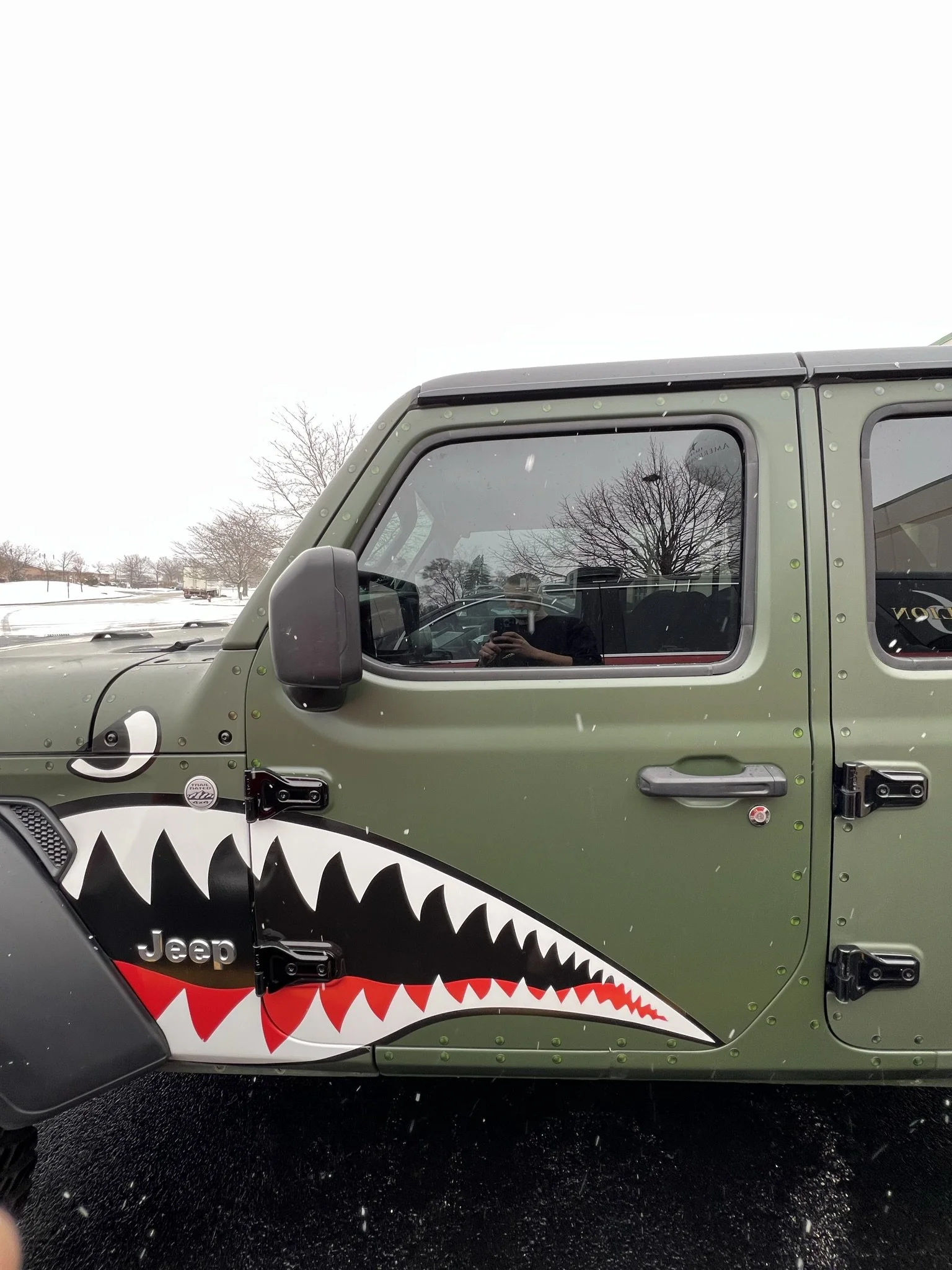 Close-up of a green Jeep with shark mouth graphic on the side, including white teeth and red tongue, parked on wet asphalt on a snowy day with leafless trees and overcast sky in the background.