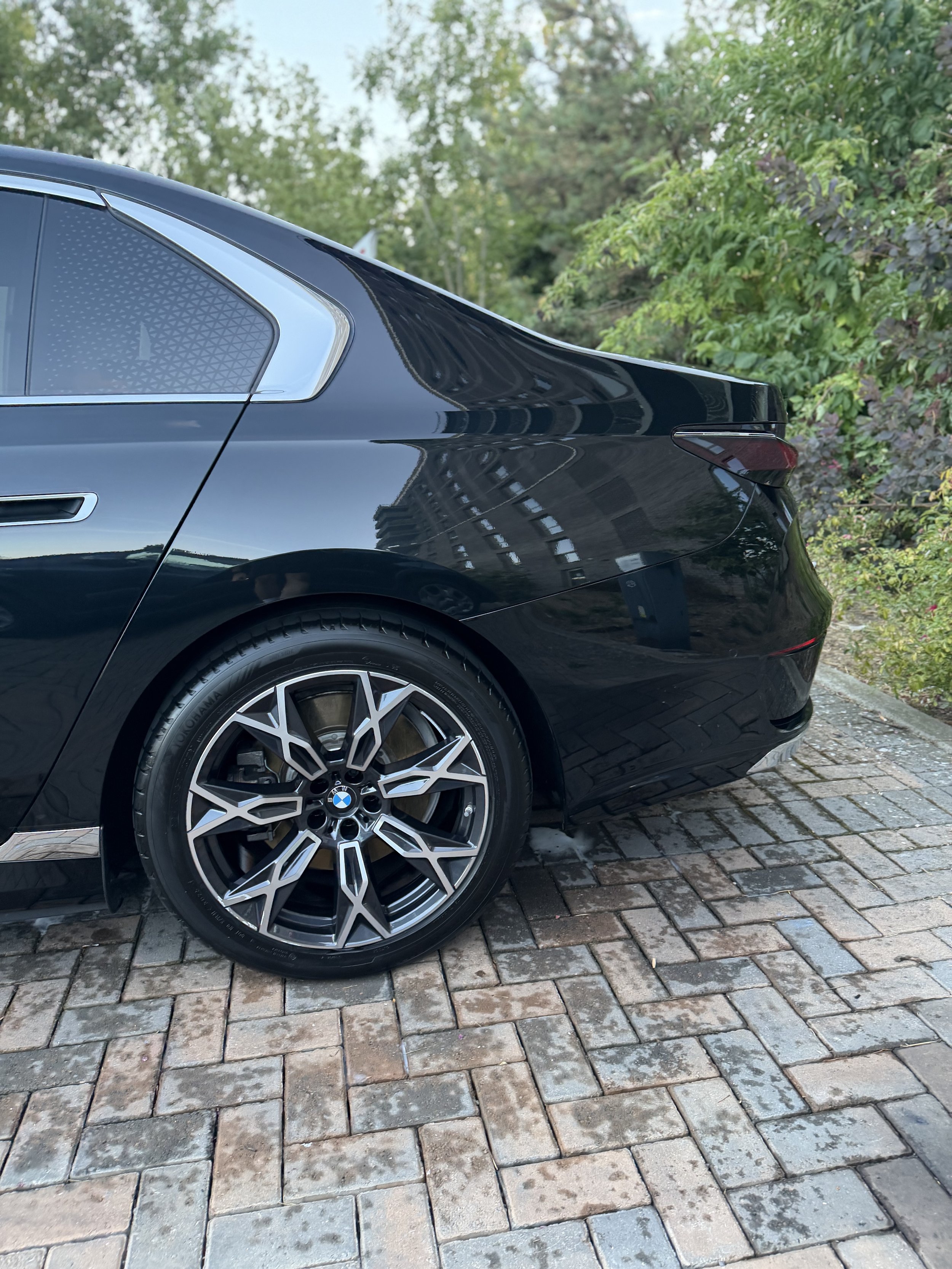 Close-up of the rear side of a shiny black BMW sedan parked on a cobblestone paved street with trees and bushes in the background.