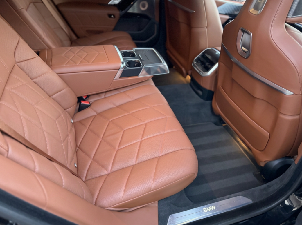 The interior of a luxury BMW vehicle showing brown leather seats, quilted with diamond stitching, with a fold-down armrest and cupholders in the center, and black carpeted floor mats.