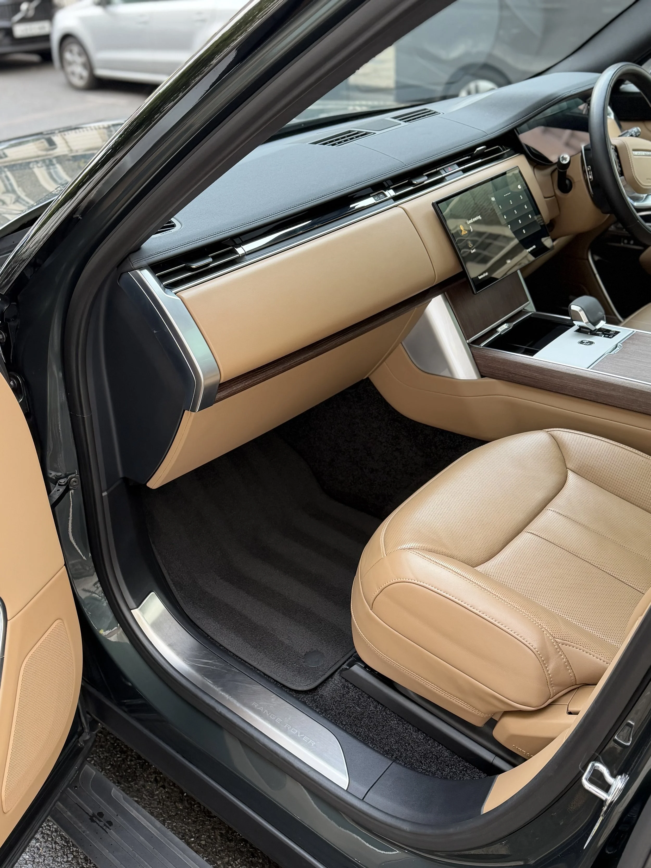 Interior of a luxury Range Rover vehicle showing a beige leather seat, a digital dashboard screen, and wood and metal accents on the dashboard and door.